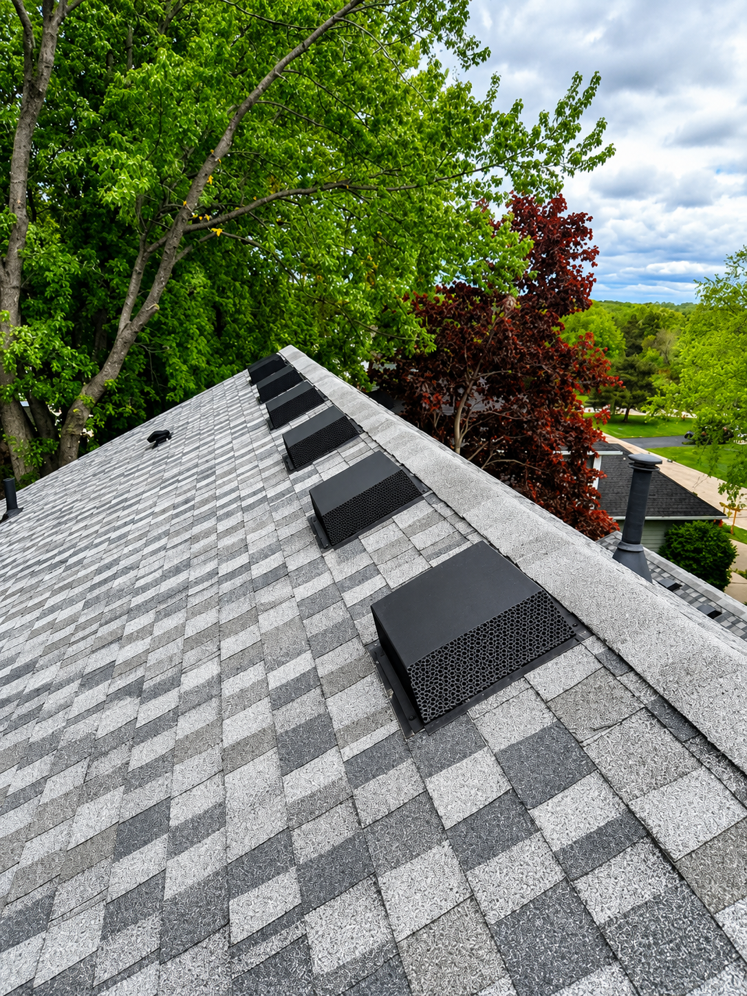 Roof with gray shingles and black vent caps beside leafy green trees and a cloudy sky