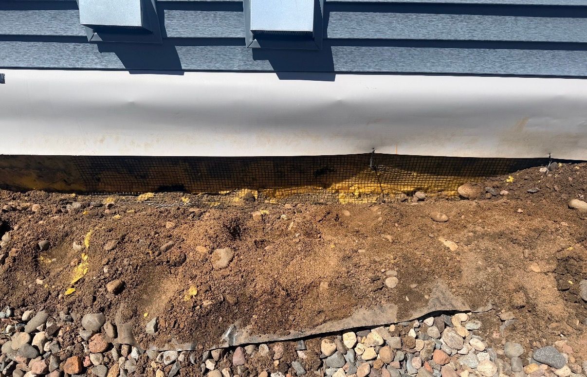Bare ground beside a house foundation with gravel and exposed soil under gray siding.