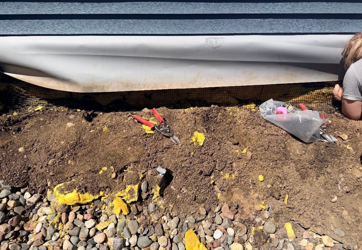 Soil bed under a house siding with scattered toy pieces and rocks, with a person partially visible at right
