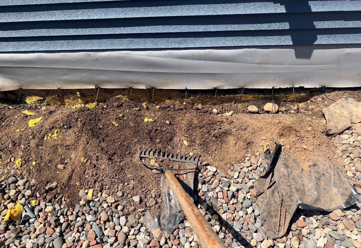 Close-up of gravel and a drainage trench beside a building wall with corrugated siding.