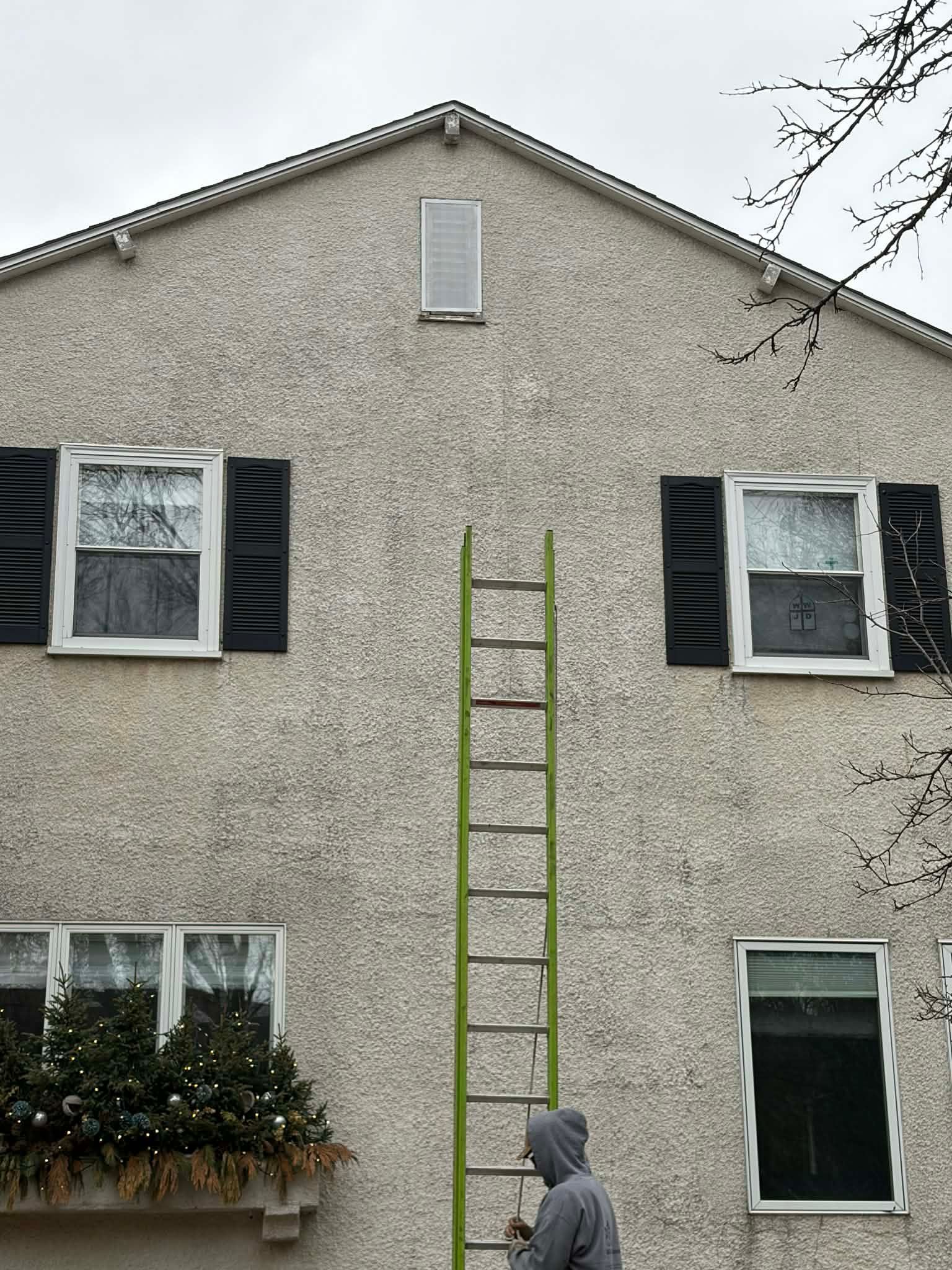 Person on a tall ladder painting a stucco house exterior with black shutters and windows