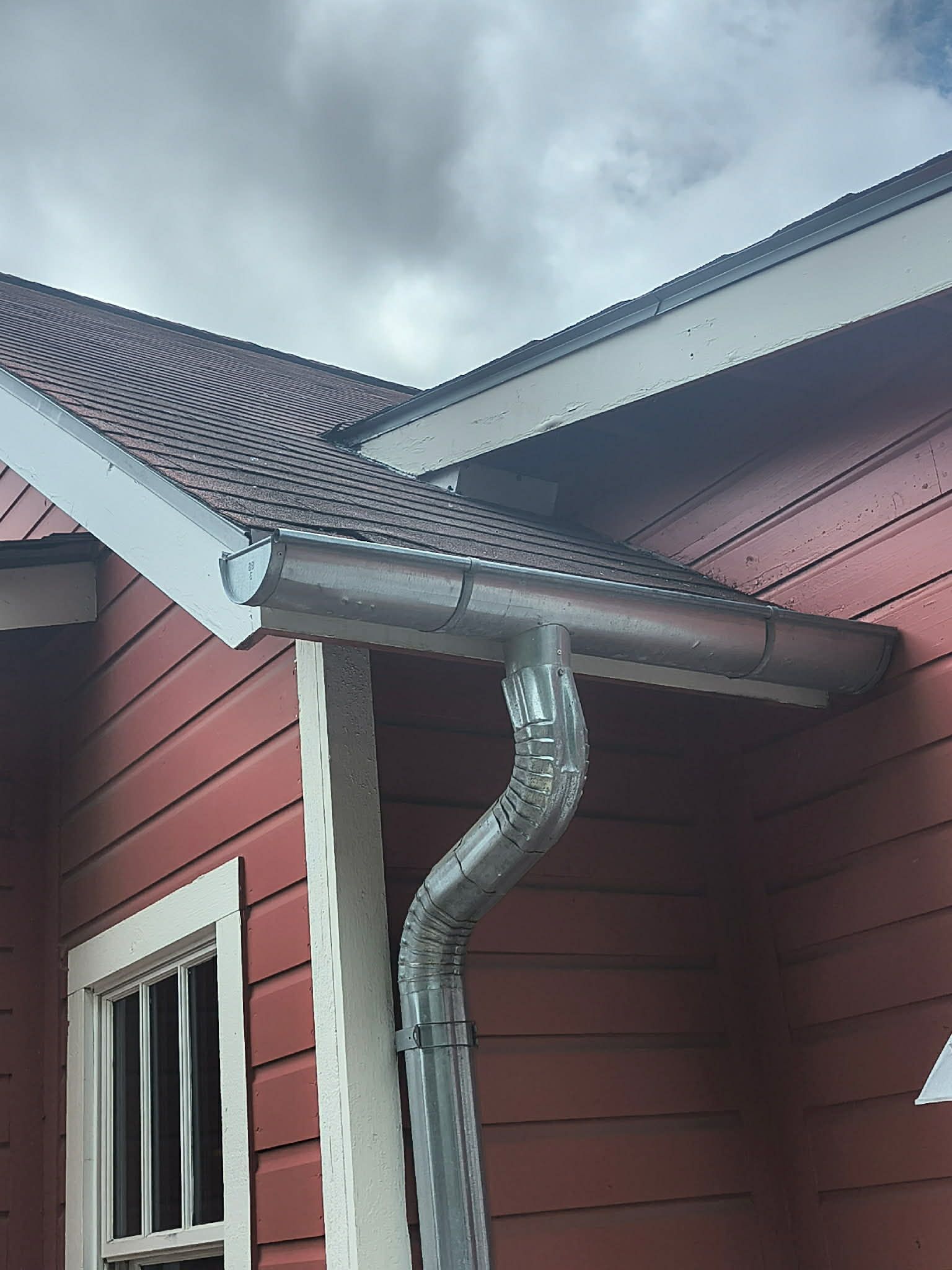 A house corner with red siding, white trim, a gray roof, and a silver downspout against a cloudy sky