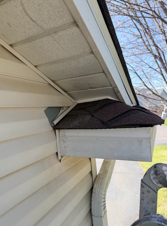 Birdhouse mounted under a house eave beside beige siding, with dark shingled roof.