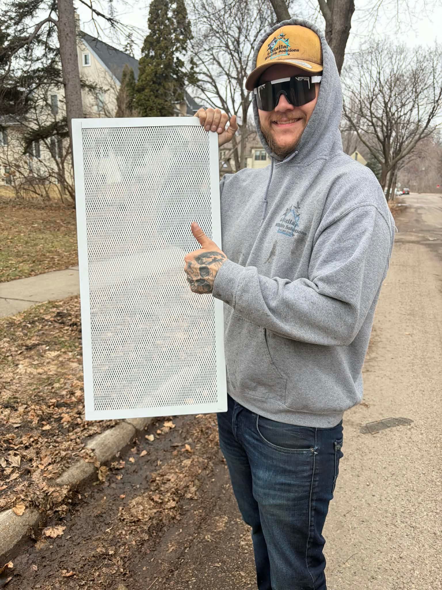 Person outdoors holding a white vent filter, wearing a gray hoodie and yellow cap near bare trees.