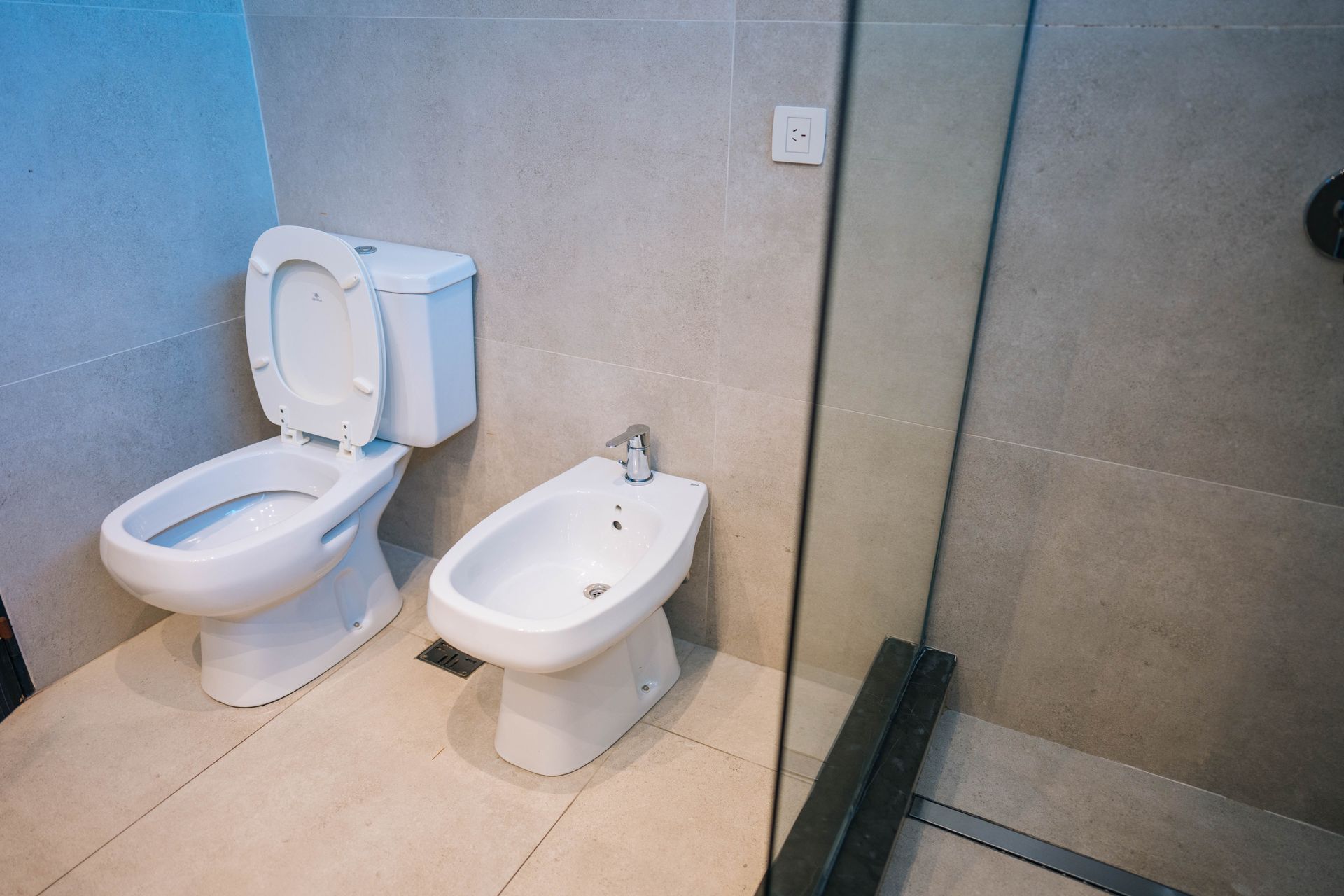 Un baño con inodoro, bidé y puerta de ducha de cristal, todo ello sobre paredes de color gris claro.