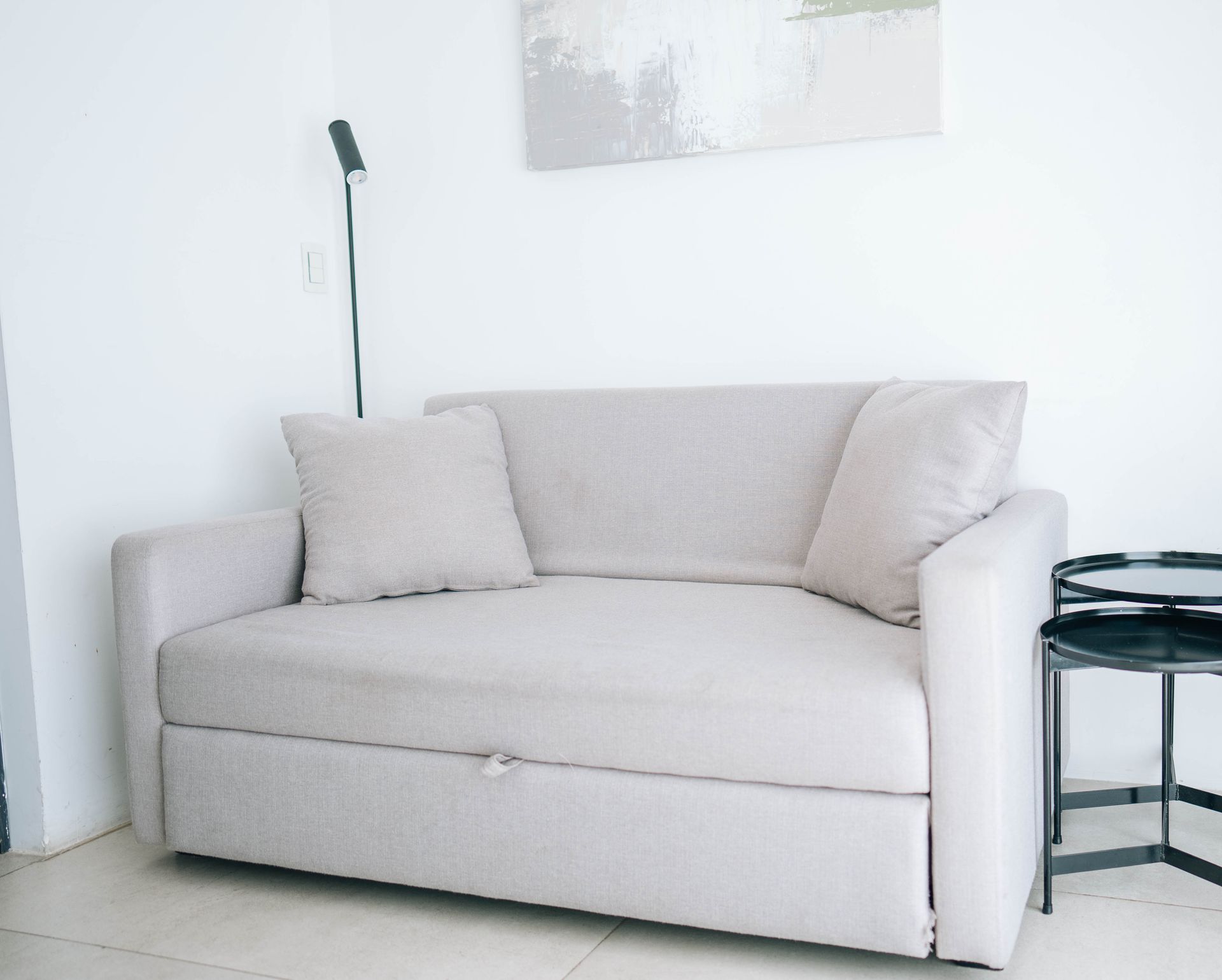 Sofá beige con dos almohadas contra pared blanca, mesa auxiliar y lámpara.