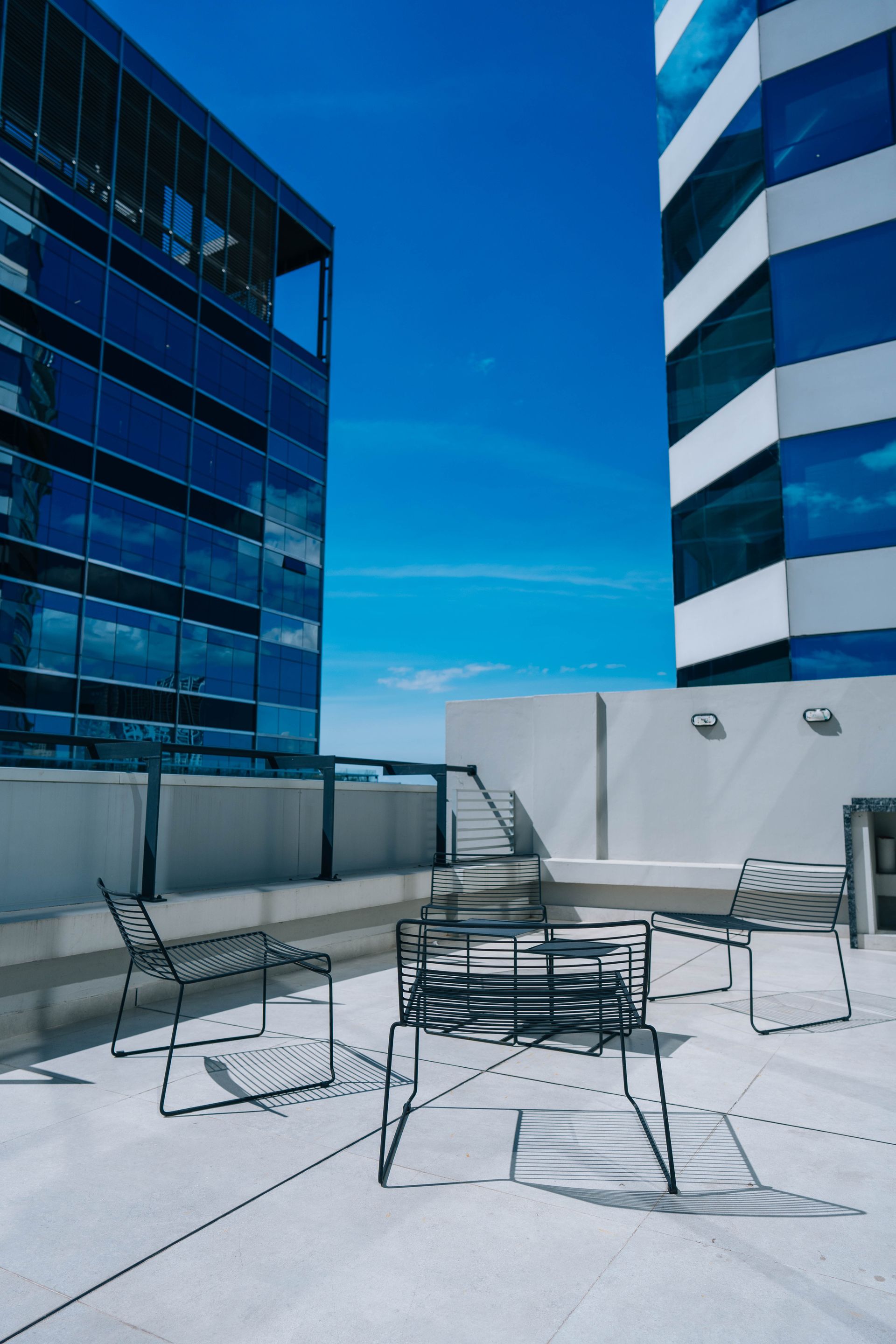 Terraza moderna: sillas de metal, mesa entre dos edificios altos con fachada de cristal y cielo azul despejado.
