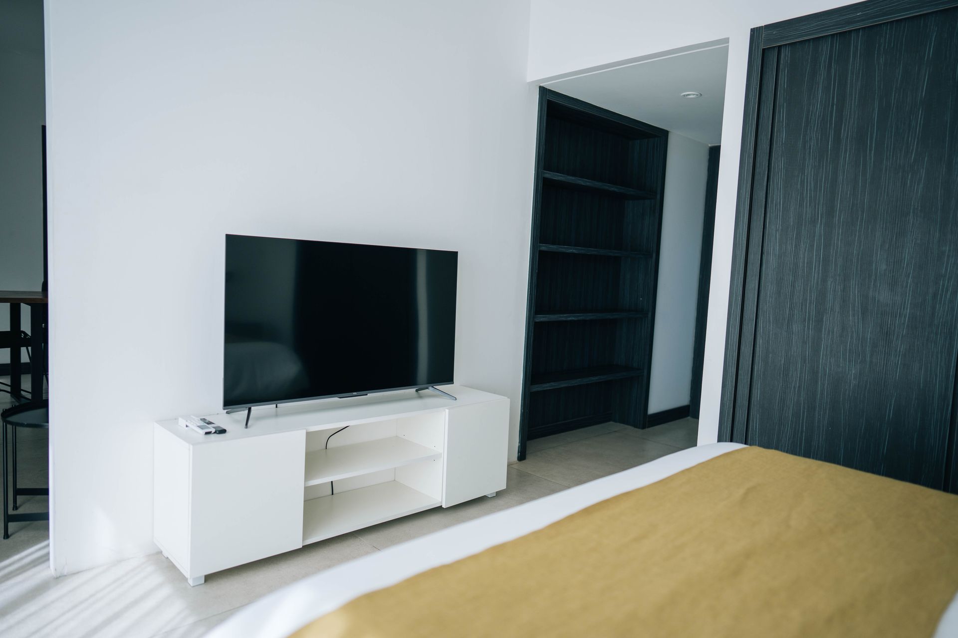Un dormitorio con un televisor de pantalla plana sobre un soporte blanco, una colcha amarilla y una estantería abierta de madera oscura.