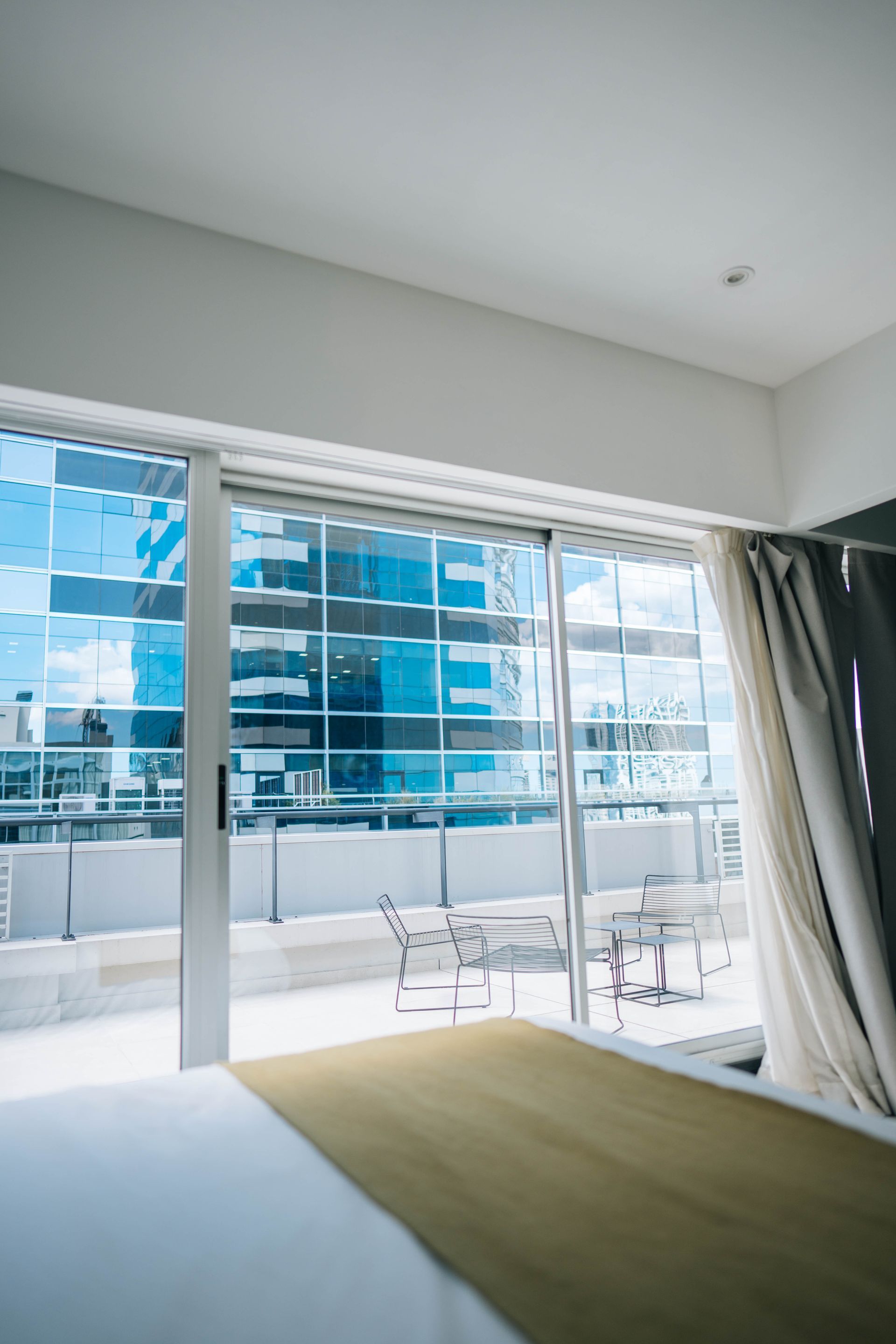 Habitación luminosa: balcón con vista a la ciudad, exterior de vidrio azul y muebles de patio de metal.
