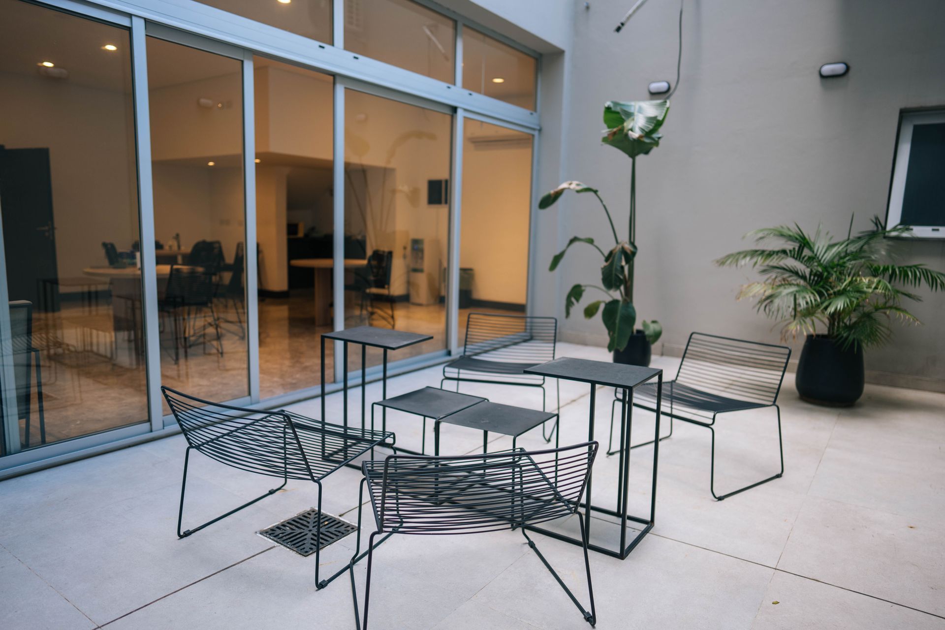 Patio moderno con sillas y mesas de metal negro sobre suelo de baldosas claras, cerca de ventanales que van del suelo al techo y plantas en macetas.