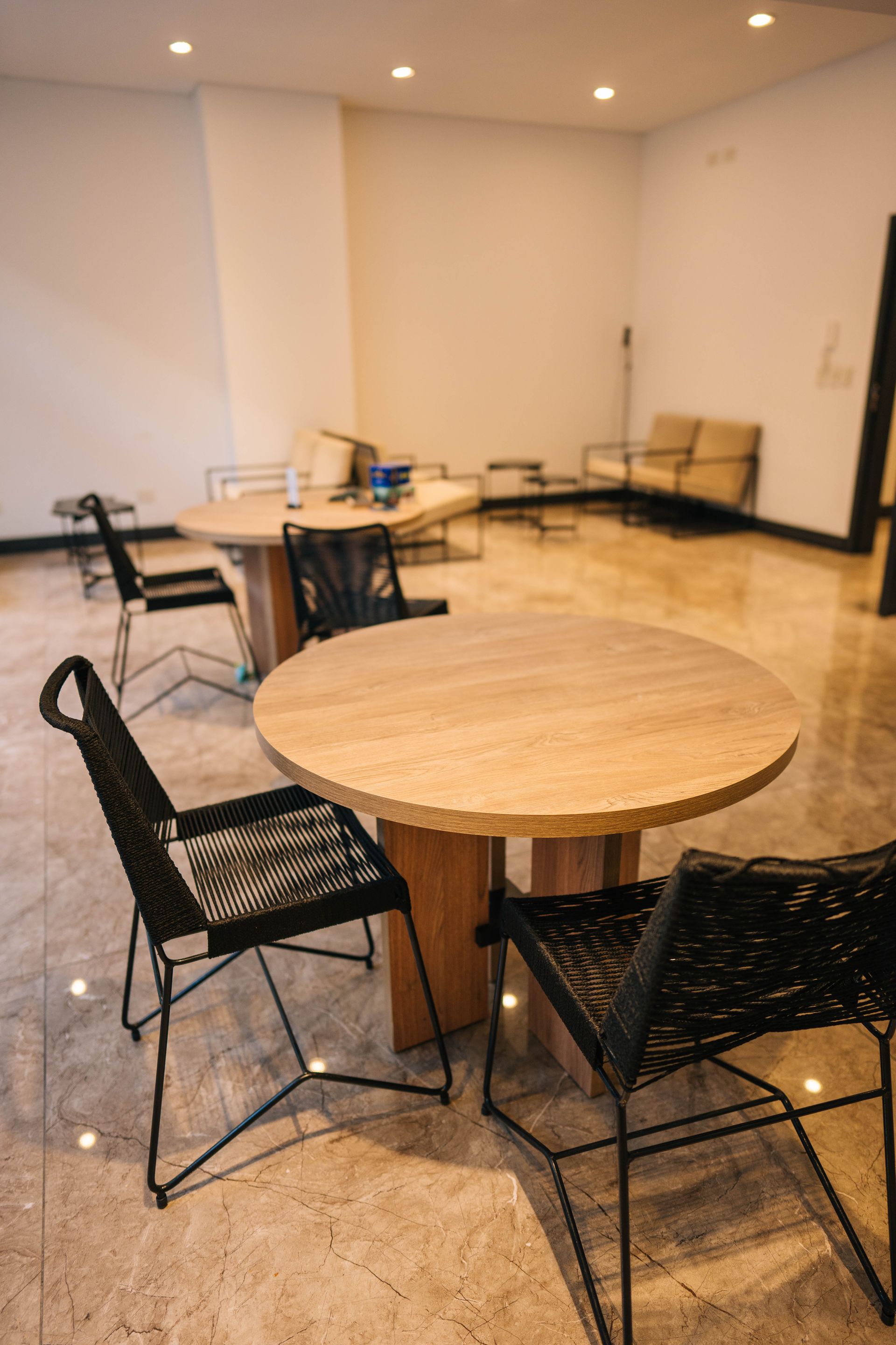 Un vestíbulo moderno con mesas de madera clara y sillas de mimbre negras sobre un suelo de baldosas pulidas, bajo luces empotradas en el techo.