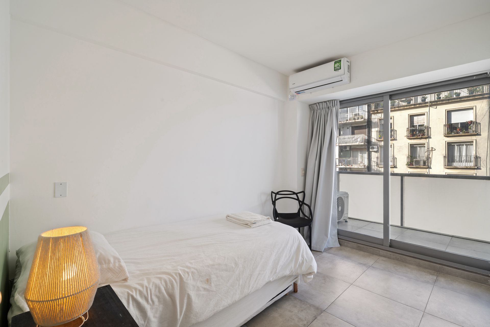 Un dormitorio minimalista de color blanco con una cama individual, una lámpara de noche, una silla oscura y una puerta corredera de cristal que da a un balcón.