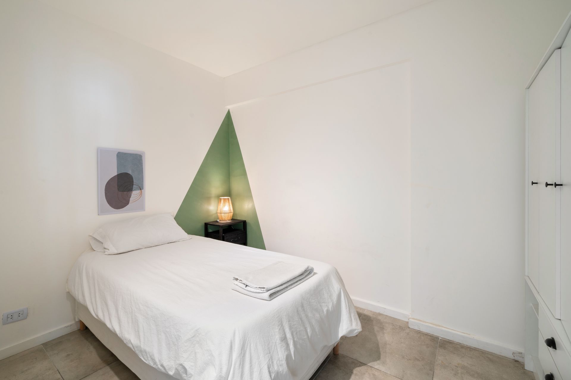 Un dormitorio pequeño y moderno con una cama individual, ropa de cama blanca, una pared decorativa triangular de color verde y una lámpara de noche.