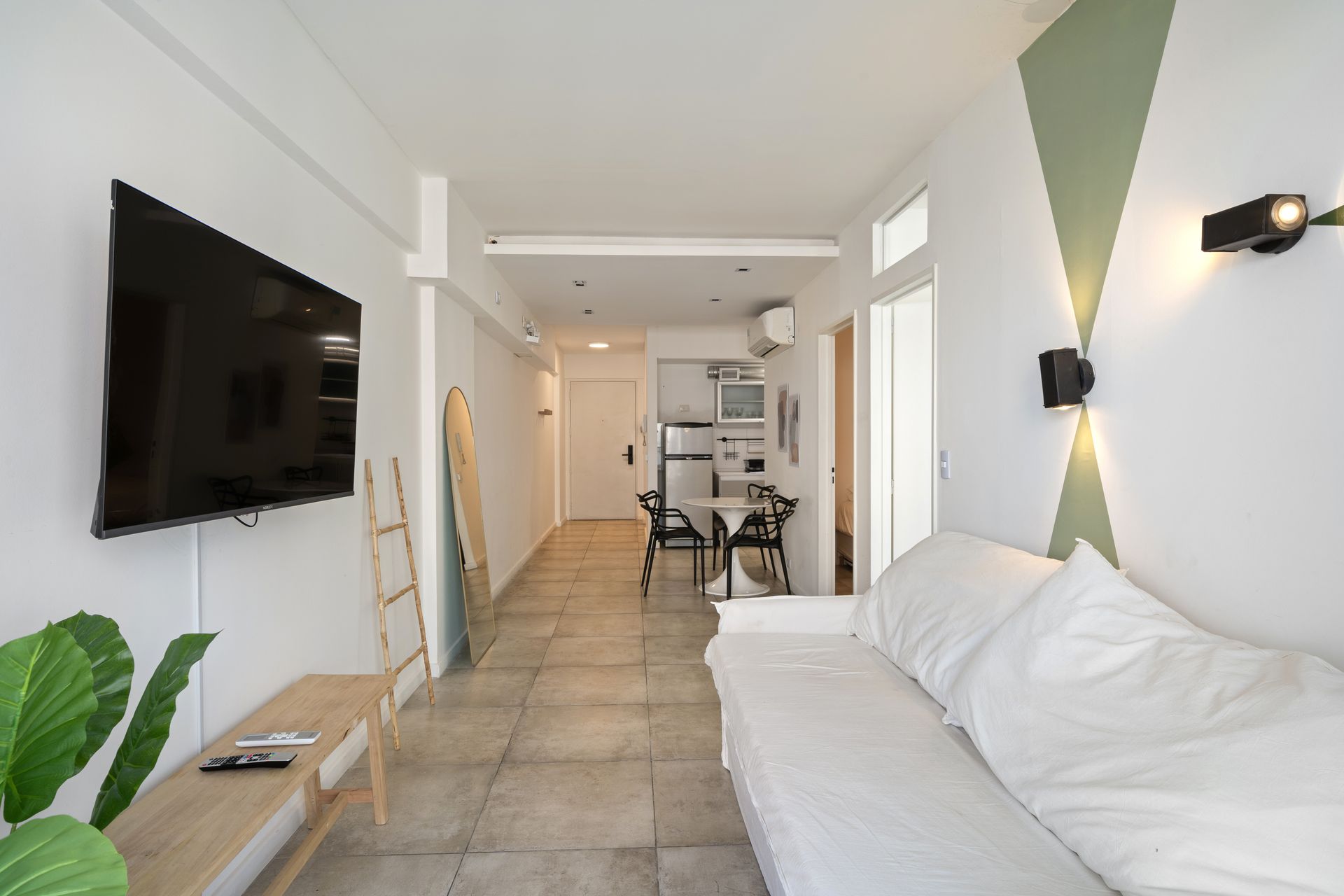 Una sala de estar tipo estudio con un sofá blanco, un televisor montado en la pared y una cocina abierta al fondo.