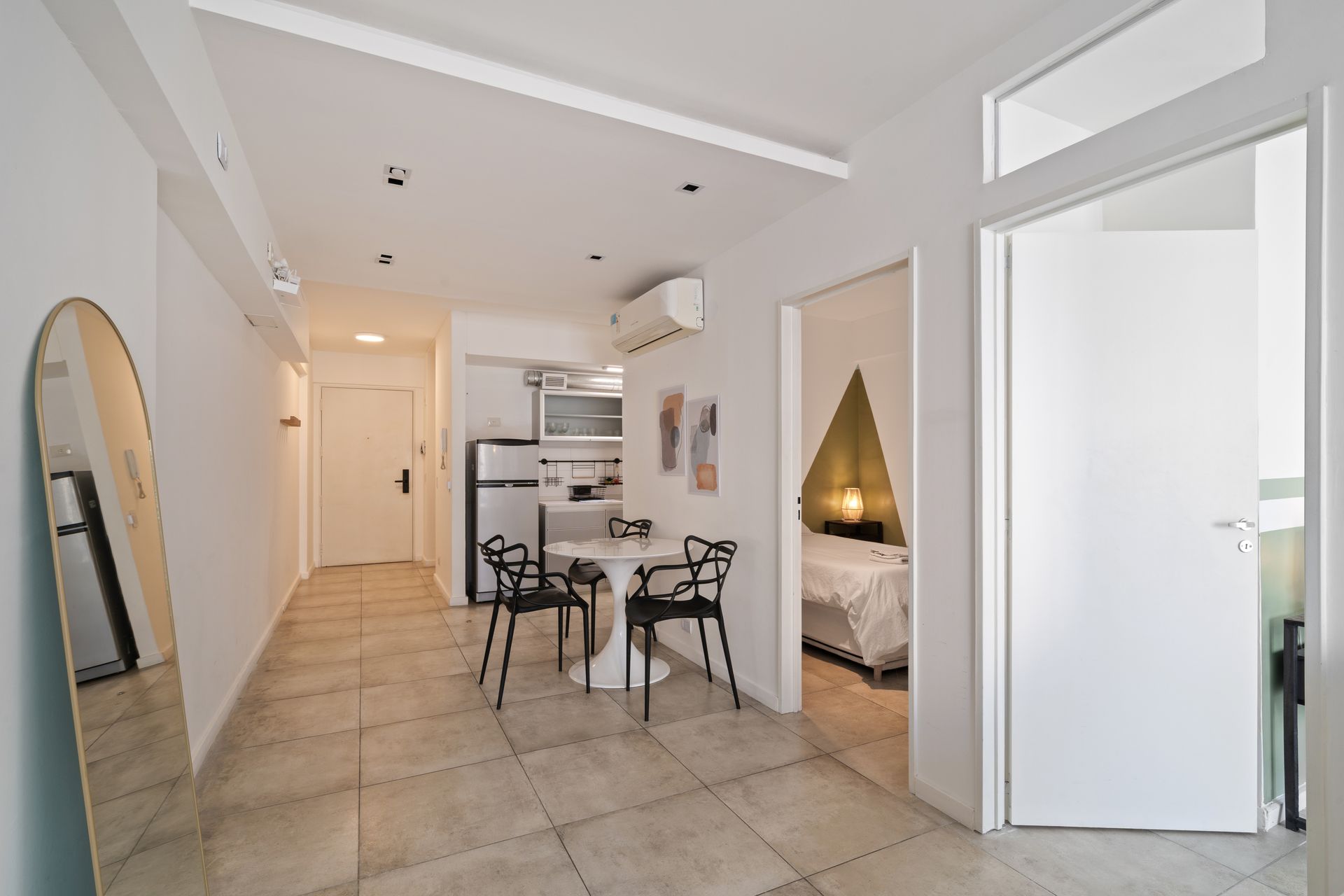 Un pequeño estudio minimalista de color blanco con una zona de cocina, una pequeña mesa de comedor y un dormitorio visible a través de una puerta.