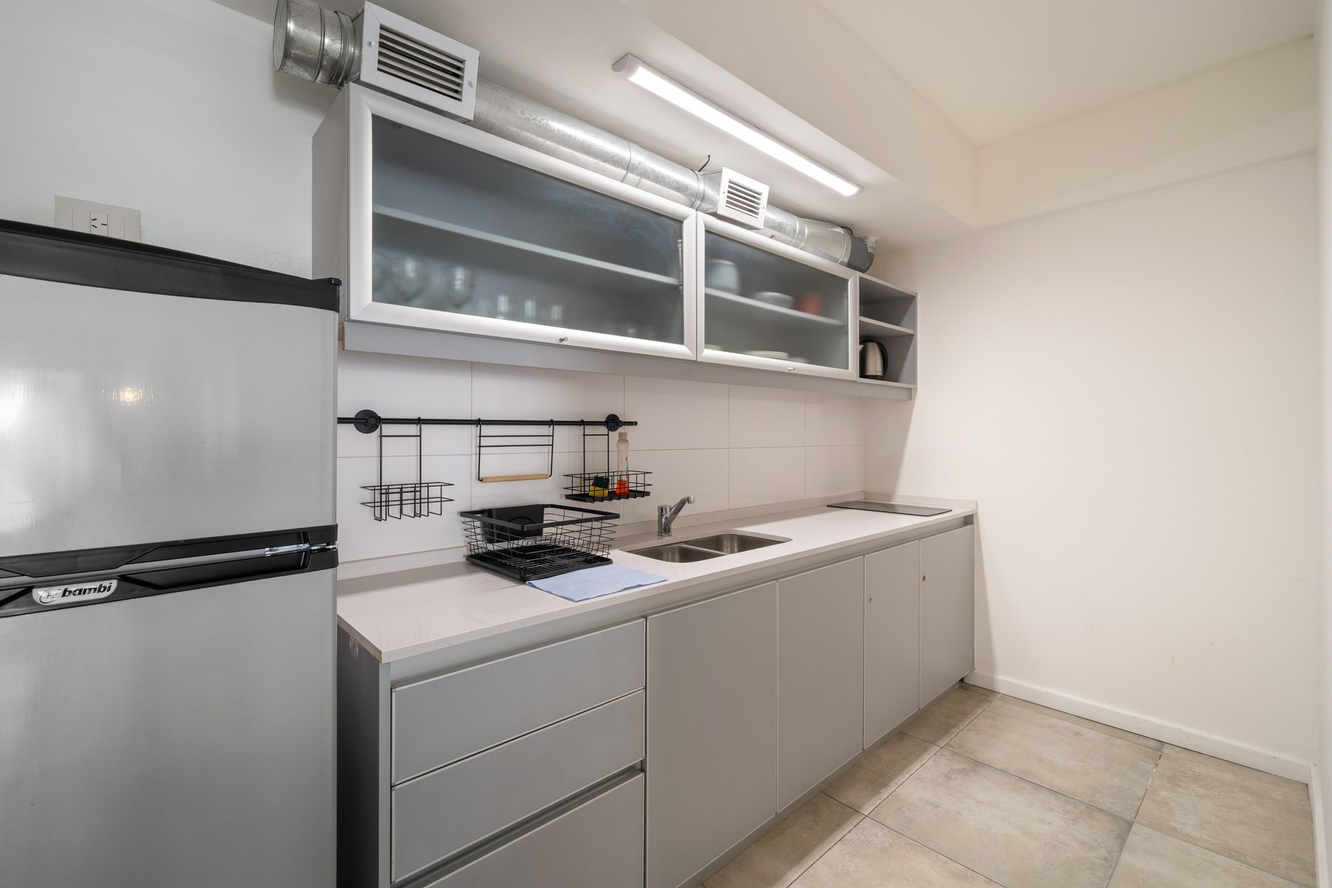 Una cocina moderna con armarios grises, un frigorífico de acero inoxidable, encimeras blancas y armarios superiores con puertas de cristal.