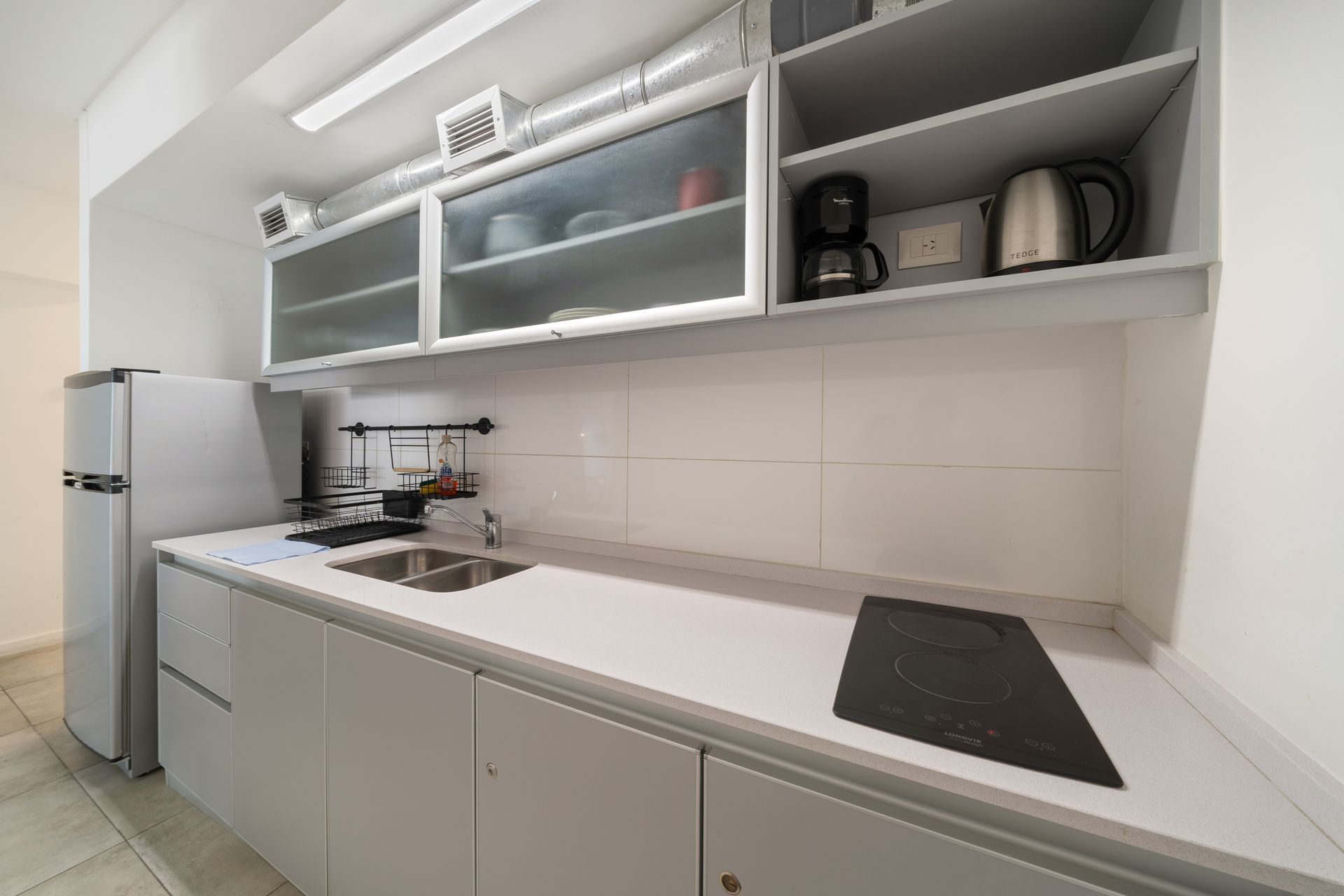 Una moderna cocina lineal de color gris claro que cuenta con refrigerador, encimera, fregadero y armarios con puertas de vidrio.