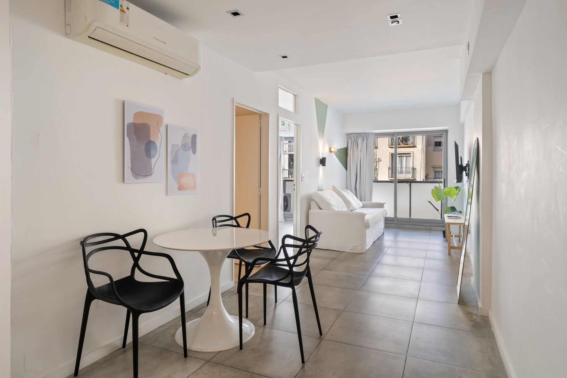 Un comedor con una mesa blanca con pedestal y dos sillas negras, que da paso a una sala de estar con sofá y balcón.