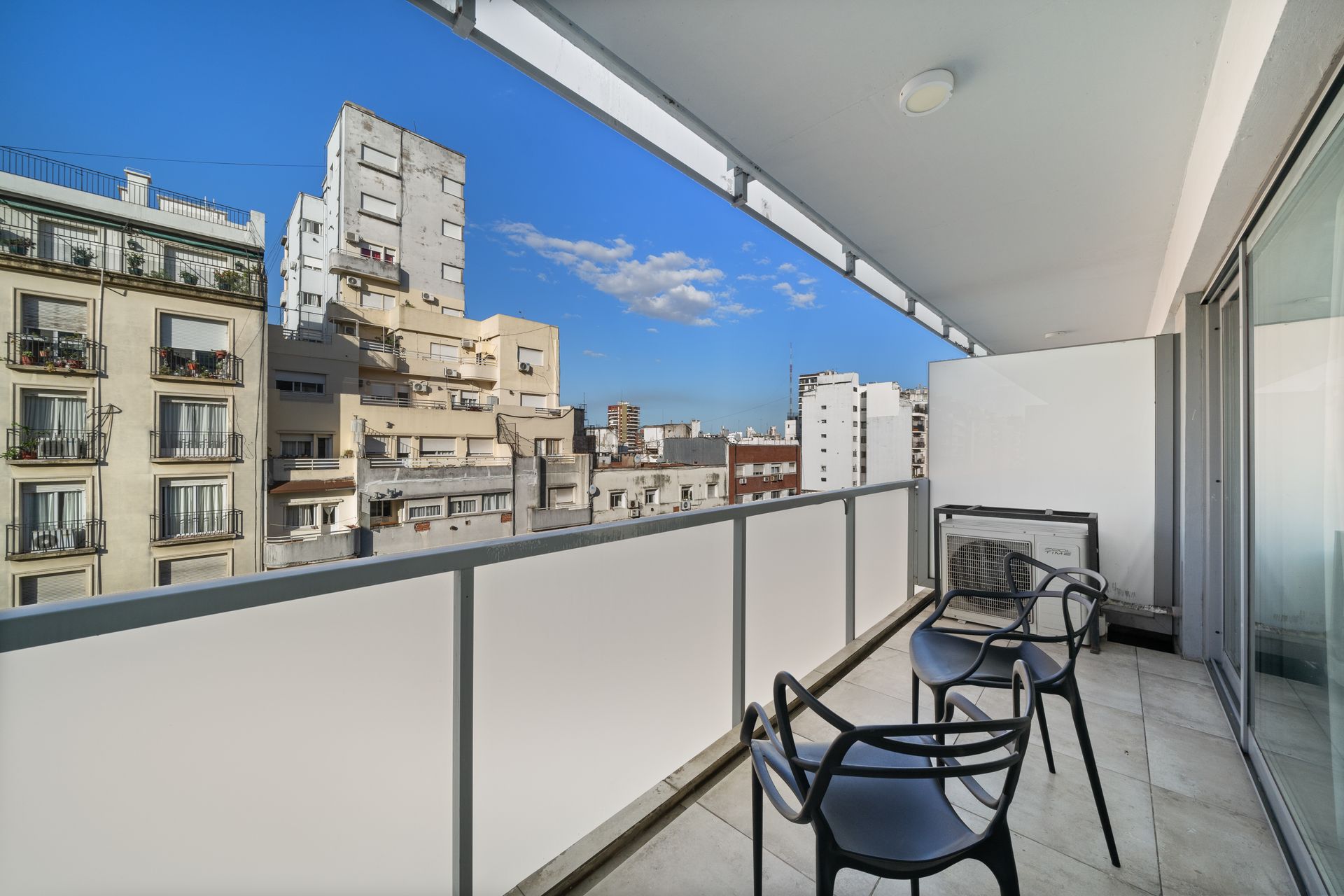 Un balcón con dos sillas oscuras con vistas al perfil urbano bajo un cielo azul despejado.