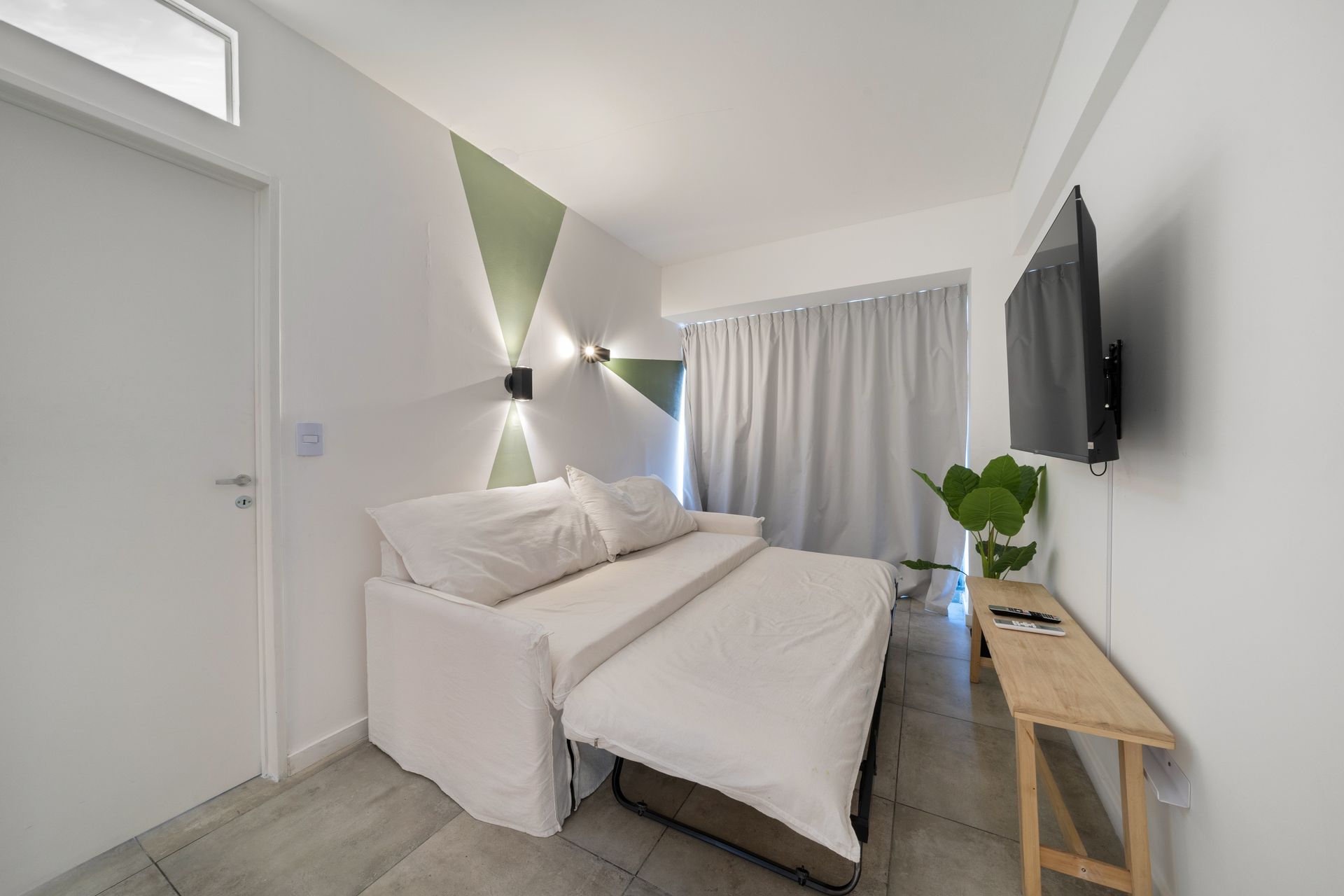 Un estudio que cuenta con un sofá cama blanco, una mesa consola de madera, un televisor montado en la pared y detalles decorativos verdes en las paredes.