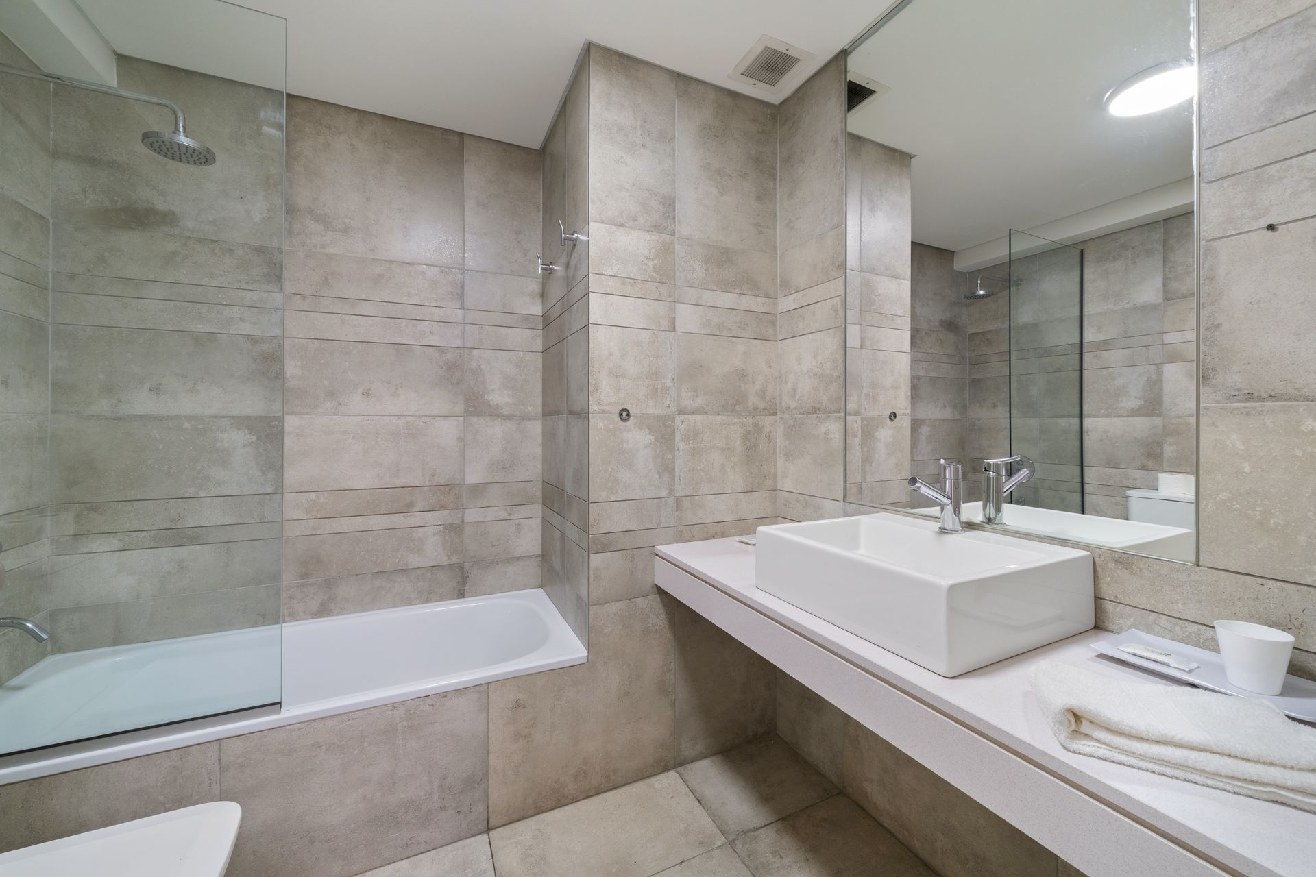 Baño moderno con paredes de piedra gris claro, bañera con mampara de ducha de cristal y lavabo blanco sobre encimera.