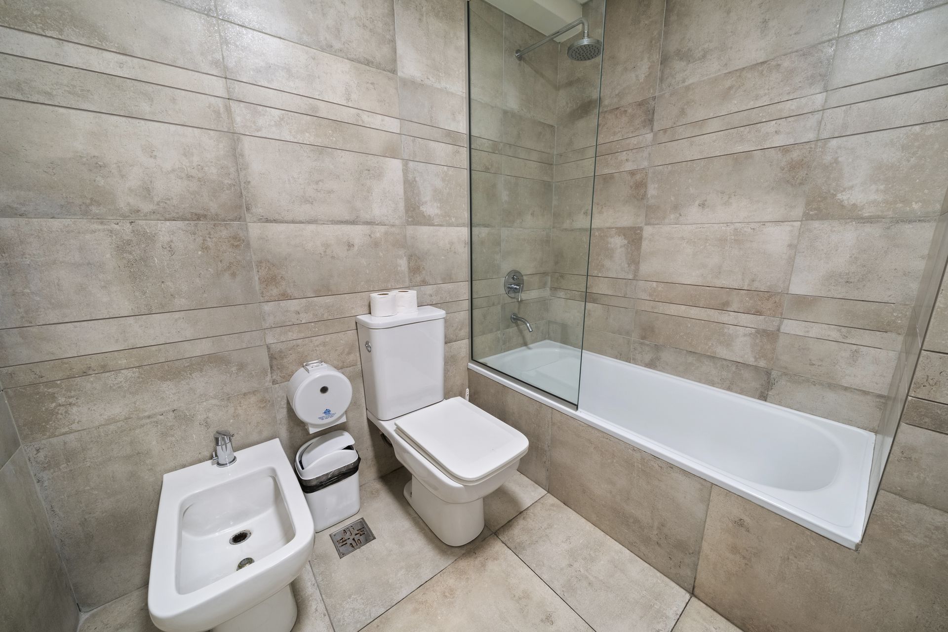 Un baño moderno con paredes alicatadas en color marrón claro, bidé, inodoro blanco y bañera con mampara de ducha de cristal.