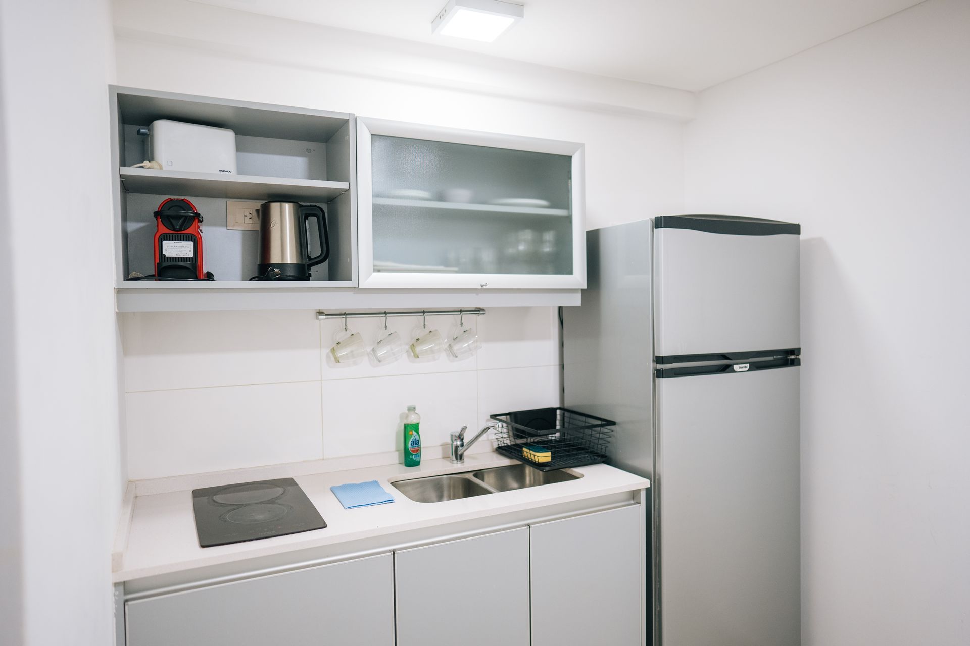 Una cocina pequeña y moderna con armarios blancos, un frigorífico plateado, un fregadero de acero inoxidable y almacenamiento montado en la pared.