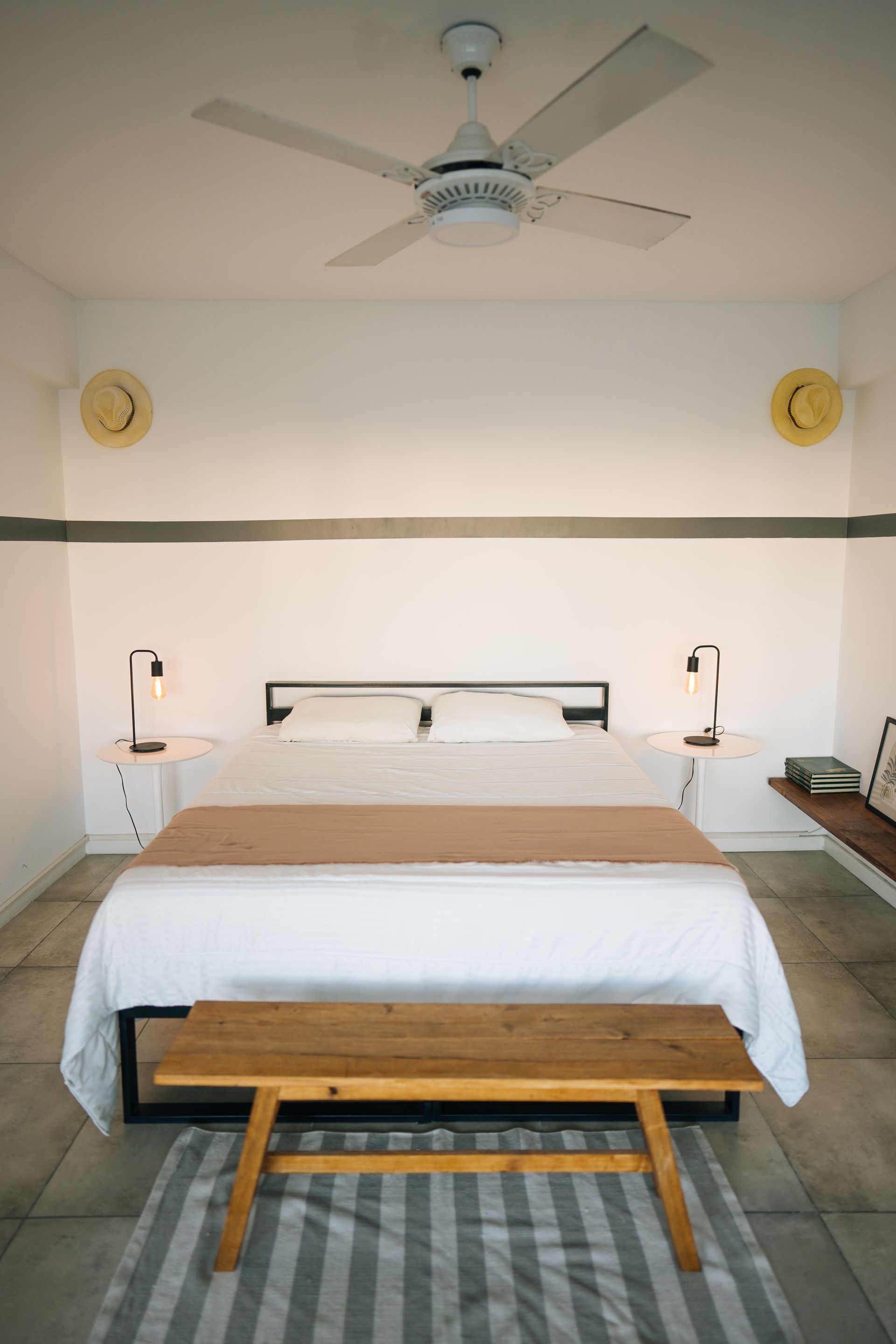 Dormitorio moderno con cama blanca, banco de madera, alfombra de rayas grises y ventilador de techo, con decoración minimalista.