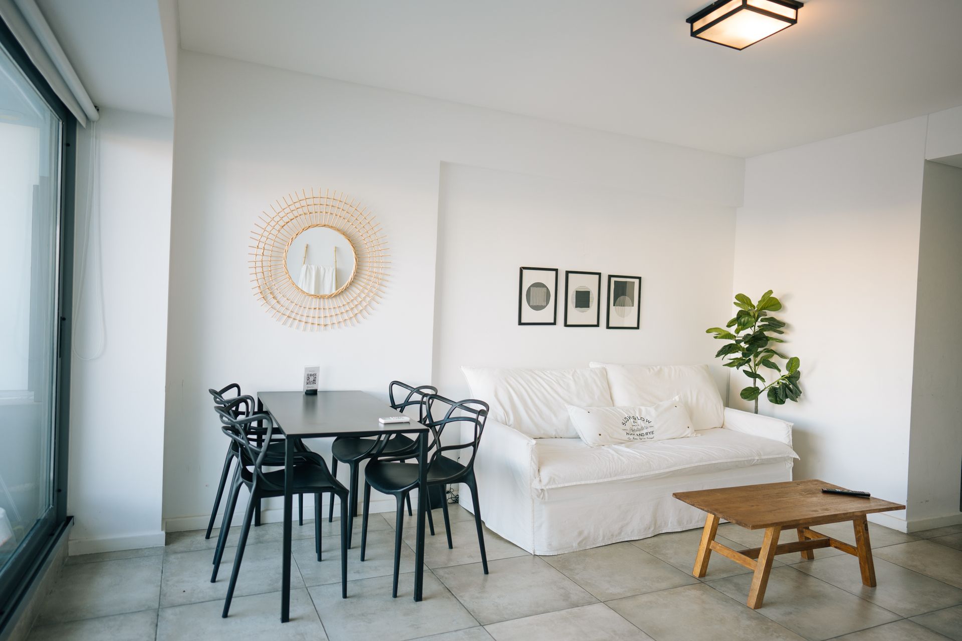 Una luminosa sala de estar y comedor con un sofá blanco, cuatro sillas negras alrededor de una mesa pequeña, una mesa de centro de madera y elementos decorativos.