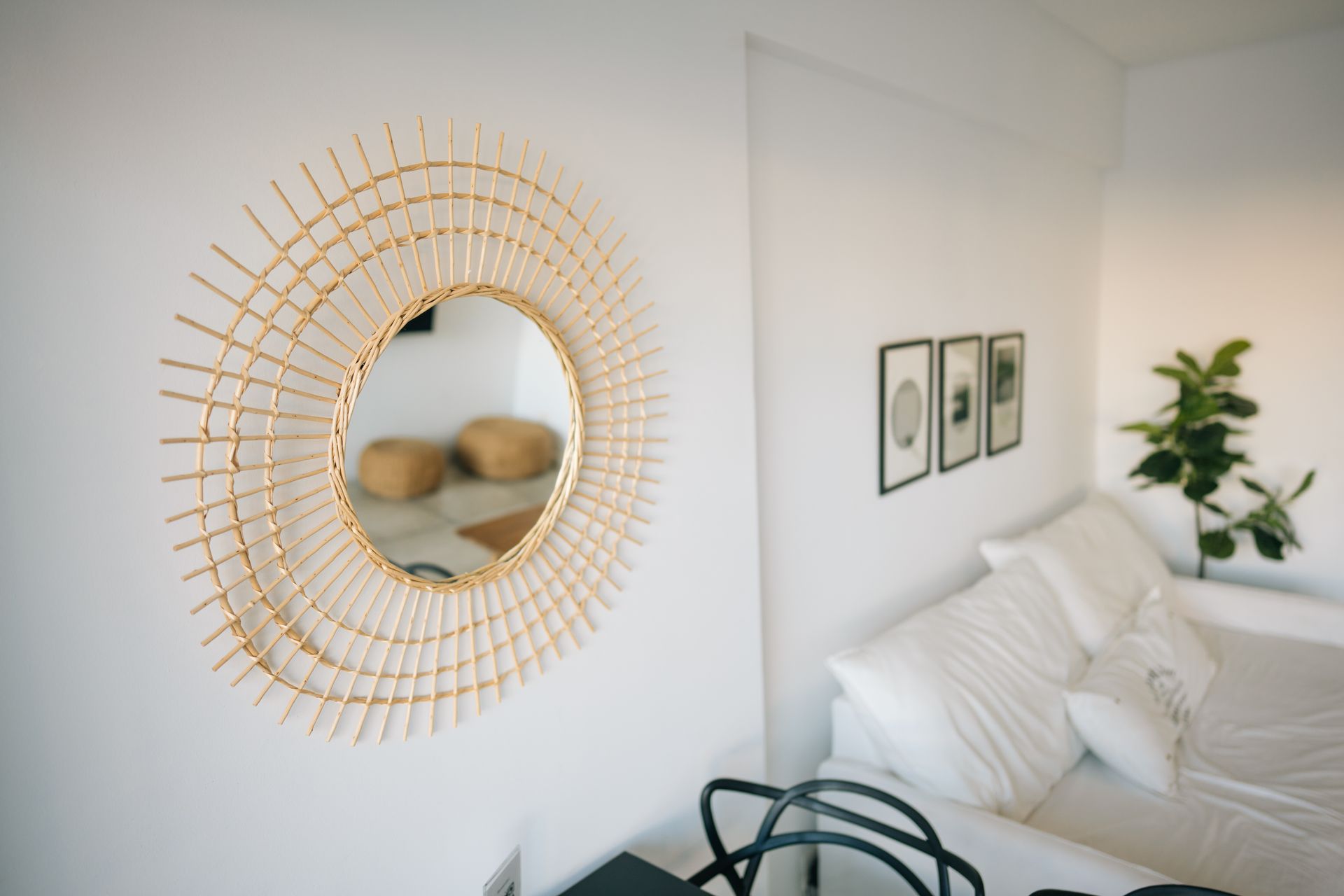Un espejo con forma de sol cuelga en una pared blanca sobre una silla de comedor, con un sofá y cuadros enmarcados visibles al fondo.