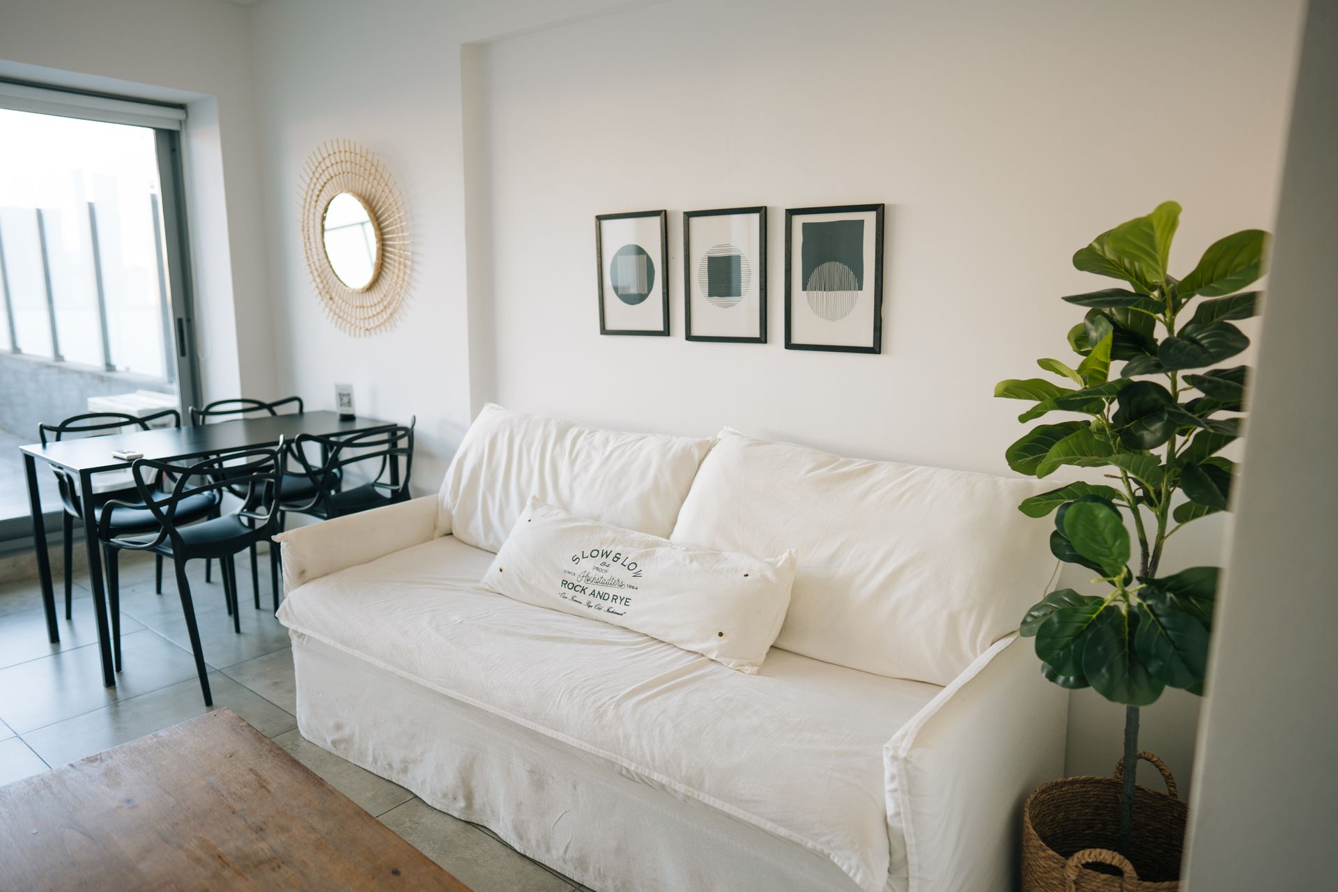 Un sofá blanco con cojín lumbar, un juego de comedor, tres láminas enmarcadas para la pared, un espejo decorativo y una planta en maceta.