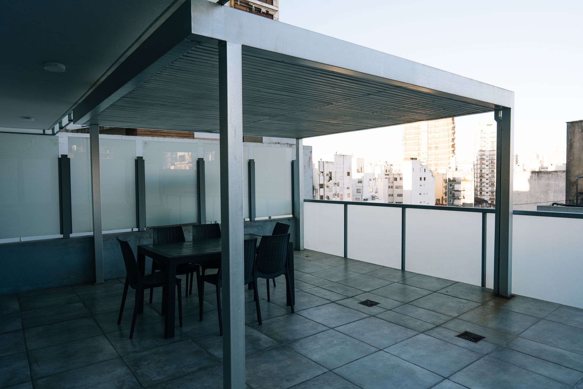 Una pérgola de metal gris da sombra a un patio exterior con zona de comedor, situado en una terraza embaldosada con vistas al perfil urbano de la ciudad.