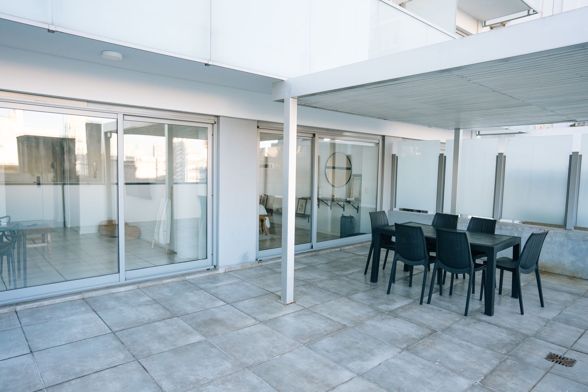 Un patio pavimentado con una pérgola blanca que cubre una mesa de comedor y sillas, situado frente a una casa moderna con puertas corredizas de vidrio.