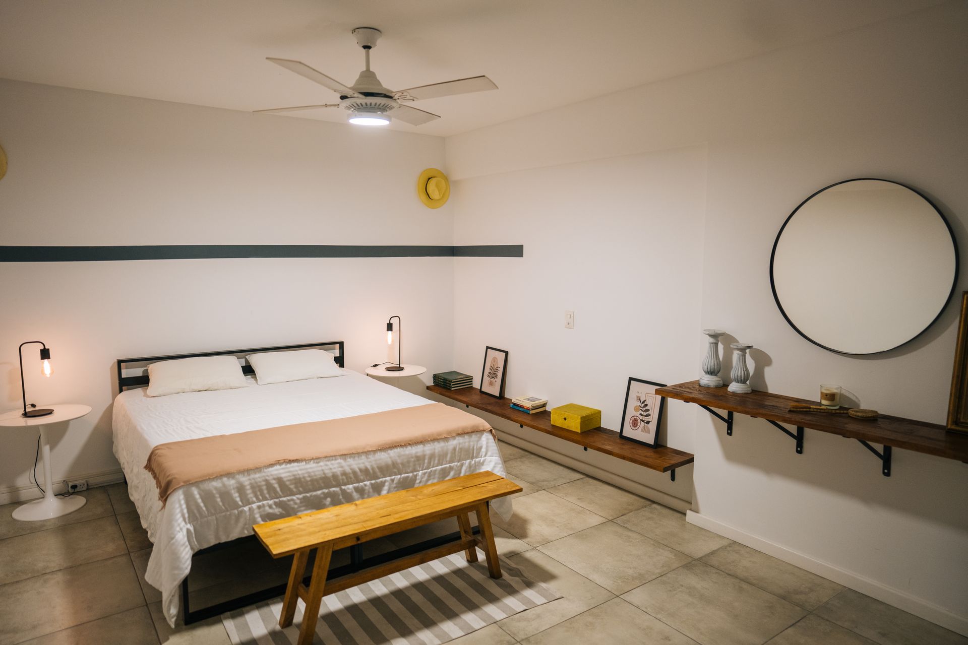 Un dormitorio minimalista con cama de matrimonio, banco de madera, estantes flotantes, espejo circular y paredes blancas con una franja azul.