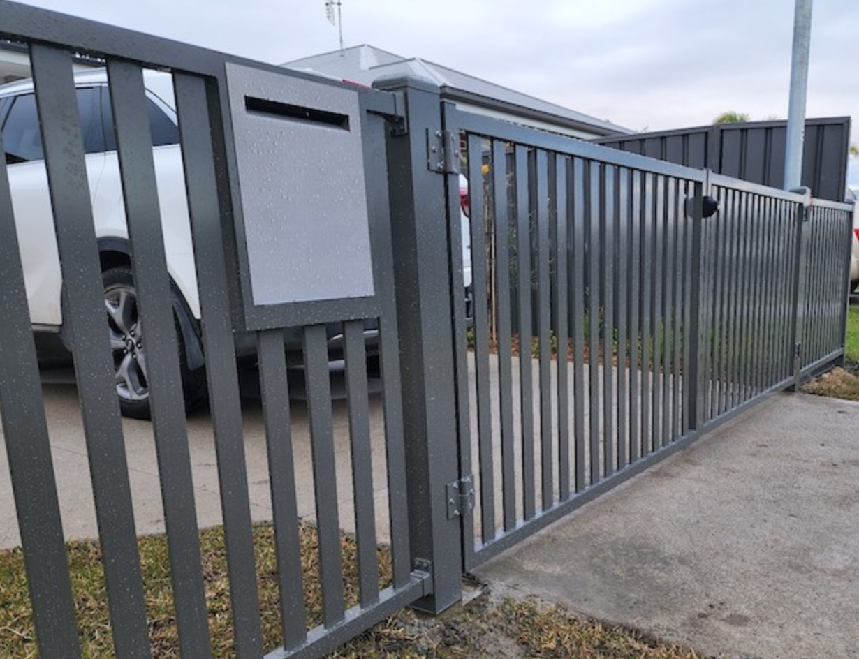 Gray metal sliding gate beside a driveway, with a white vehicle partially visible behind it.