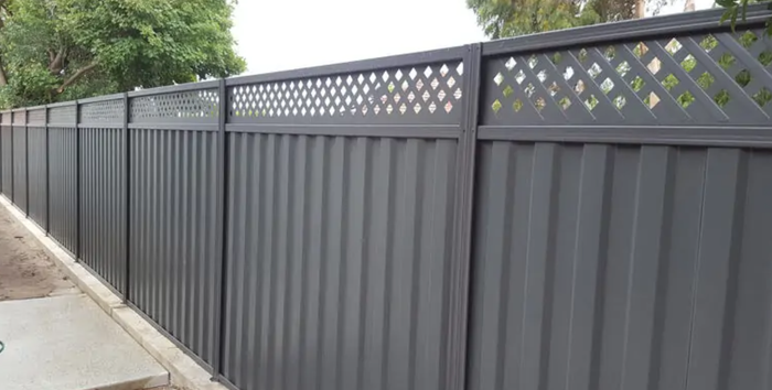 Long dark gray metal fence along a sidewalk, with lattice top panels and trees behind it.