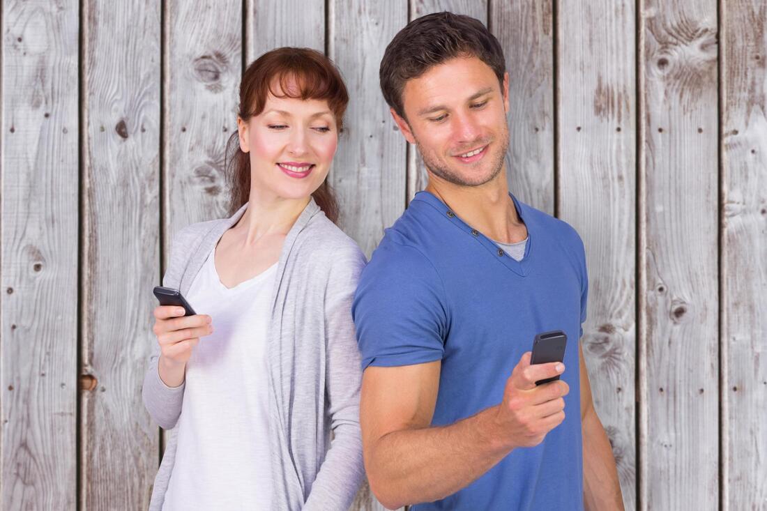 Two people standing back to back, smiling while holding smartphones against a wooden wall.