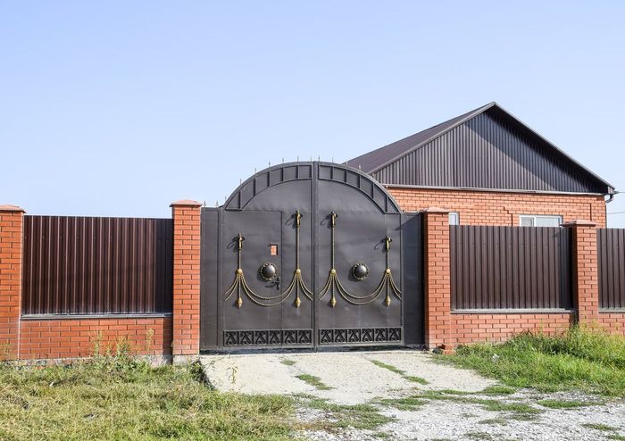 Large black metal gate in a red-brick fence, with a house and blue sky behind it