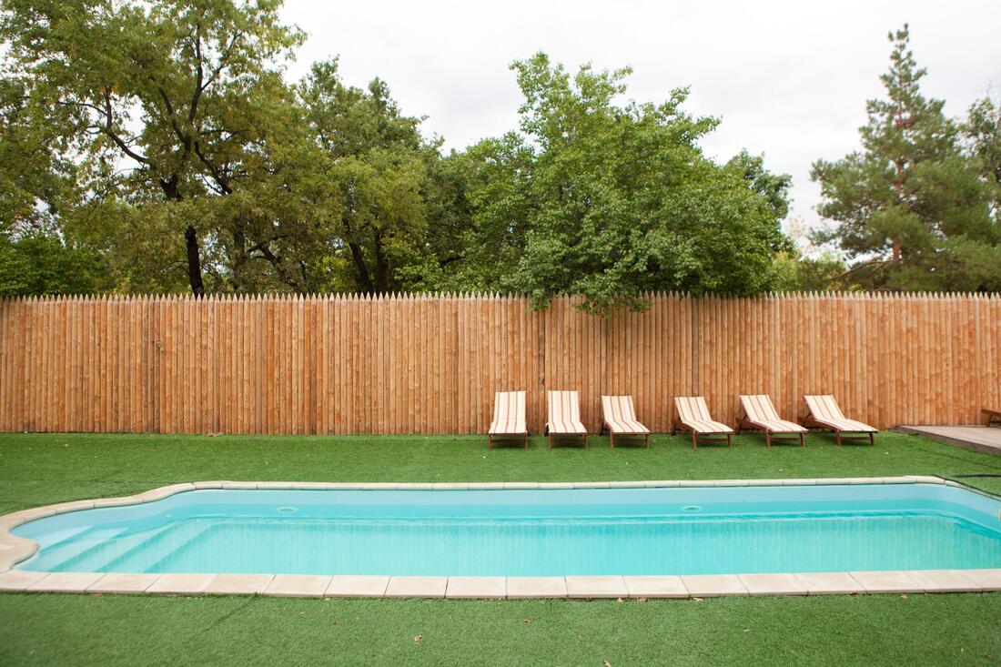 Backyard pool with lounge chairs, wooden fence, and trees behind it.