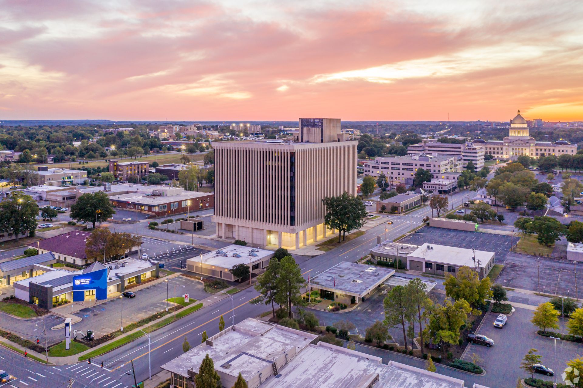 Aerial view of a downtown cityscape at sunset with midrise buildings, streets, and a warm pink sky