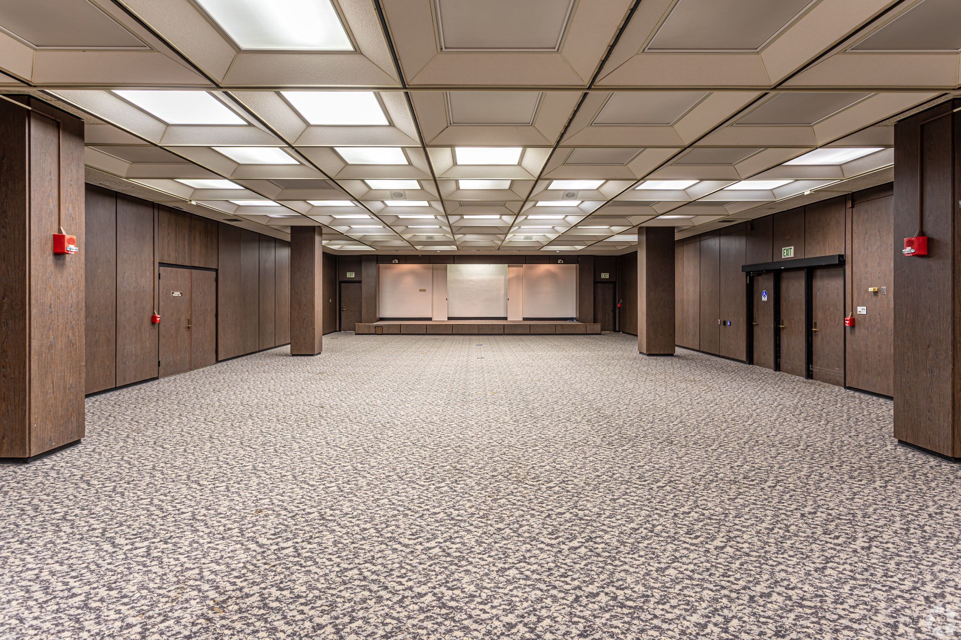 Empty carpeted conference hall with wood-paneled walls, ceiling lights, and stage at the far end