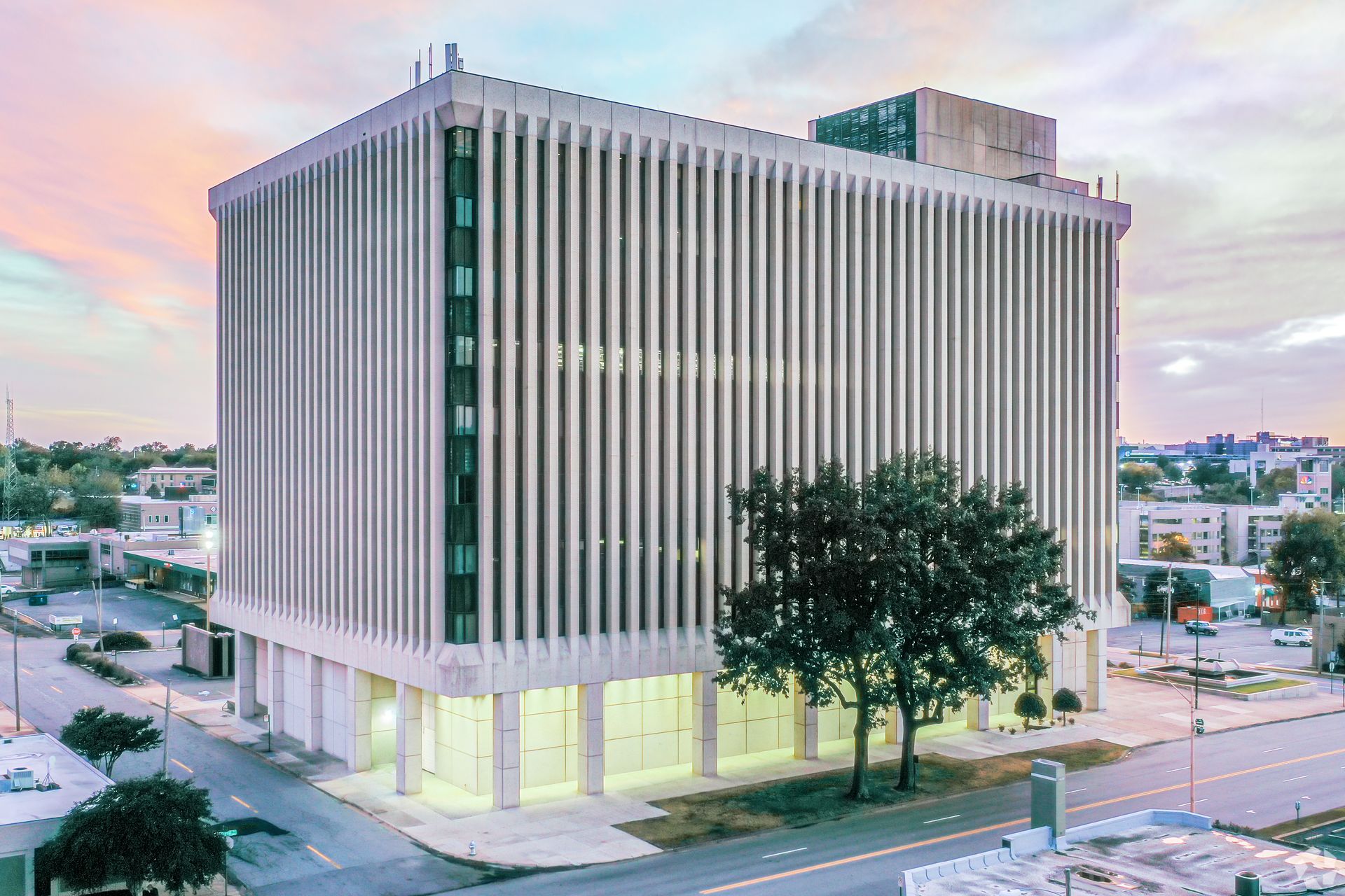 Modern office building with vertical white facade panels at dusk, viewed from a street corner with trees.