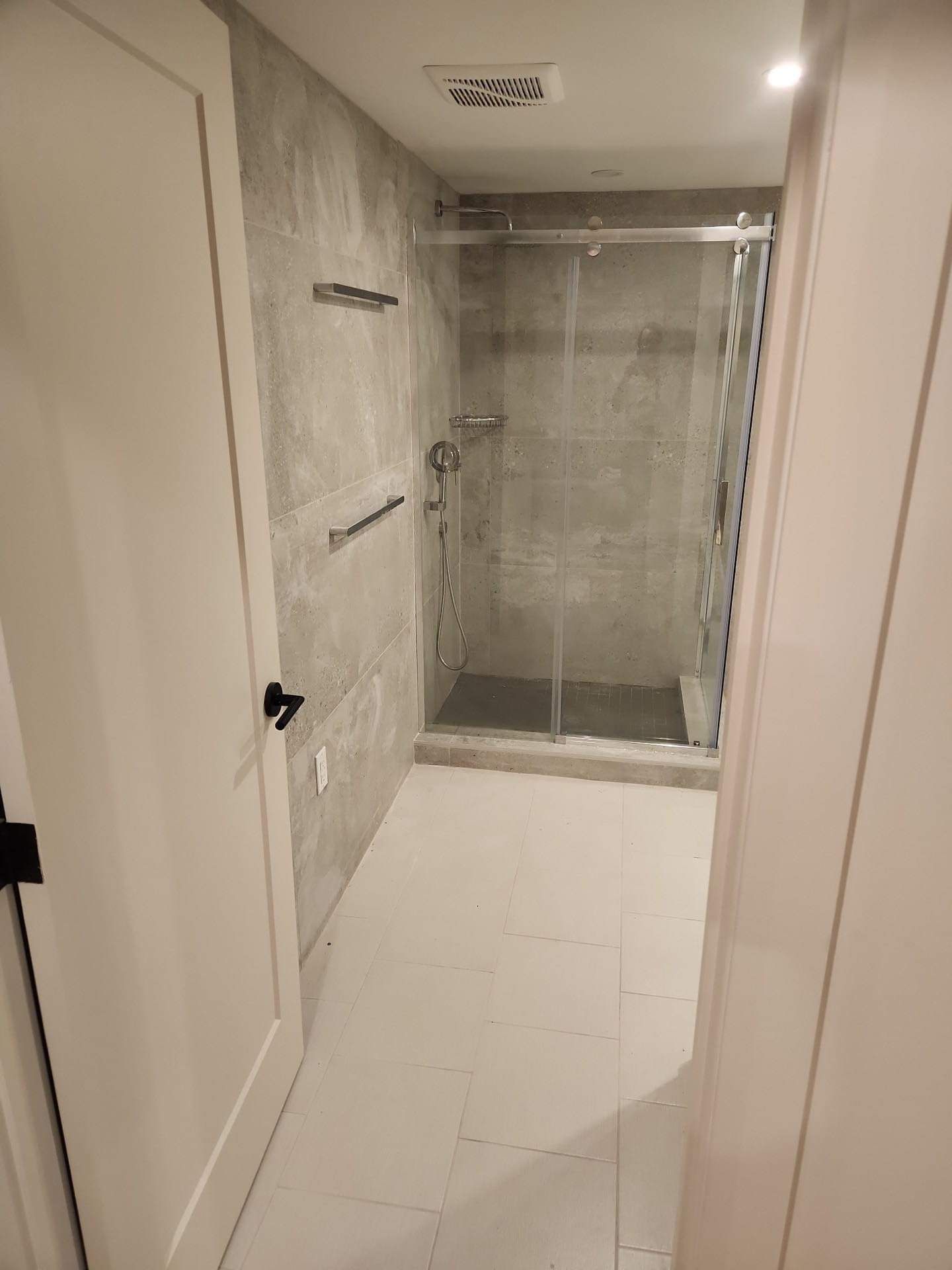 Salle de bain avec douche en verre, murs carrelés gris, sol blanc, luminaires chromés.