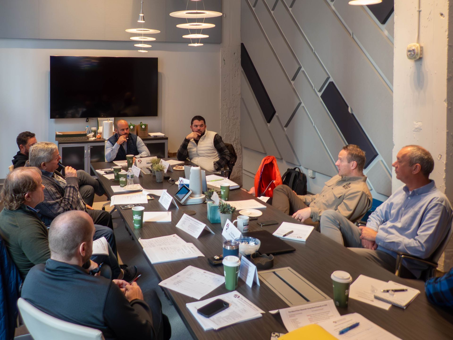 A group of people sit around a large conference table in a modern, neutral-toned office meeting room.