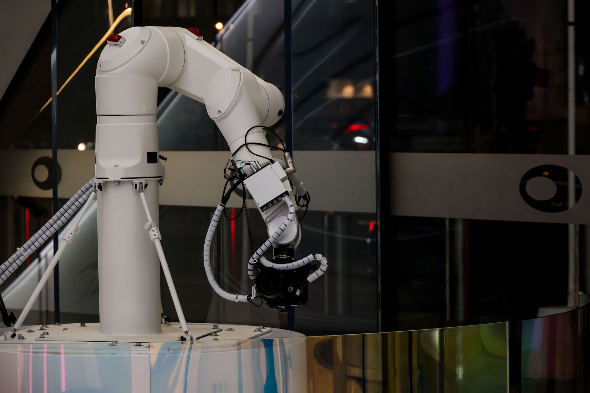 White robotic arm with a gripper in a lab or industrial setting, reaching toward a clear panel.