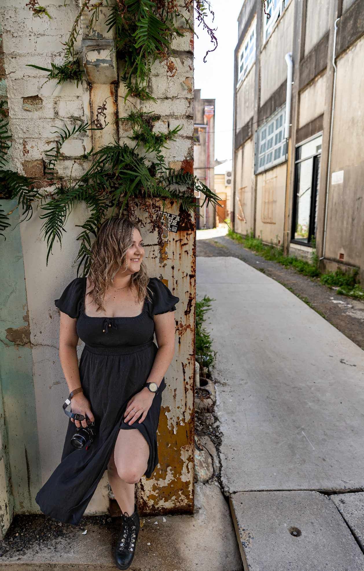 A woman in a black dress is leaning against a wall in an alleyway.