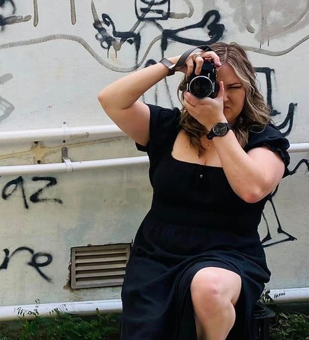 Woman in black dress taking a photo with a camera. She's outdoors near graffiti, holding the camera up to her eye.