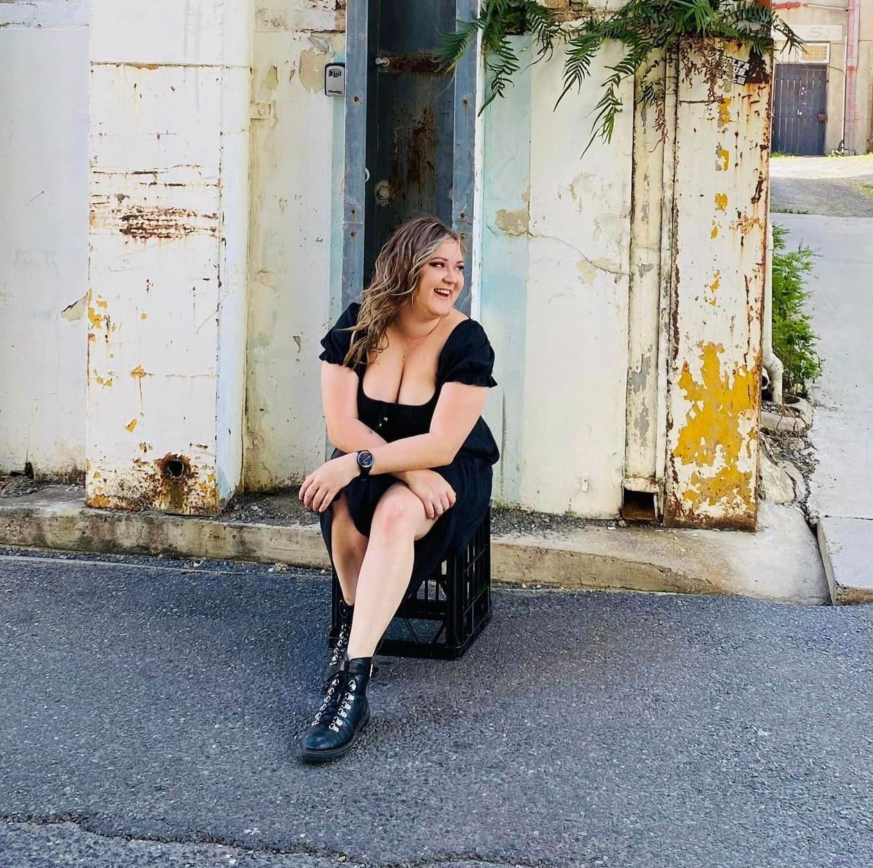A woman in a black dress is sitting on a crate in front of a building.
