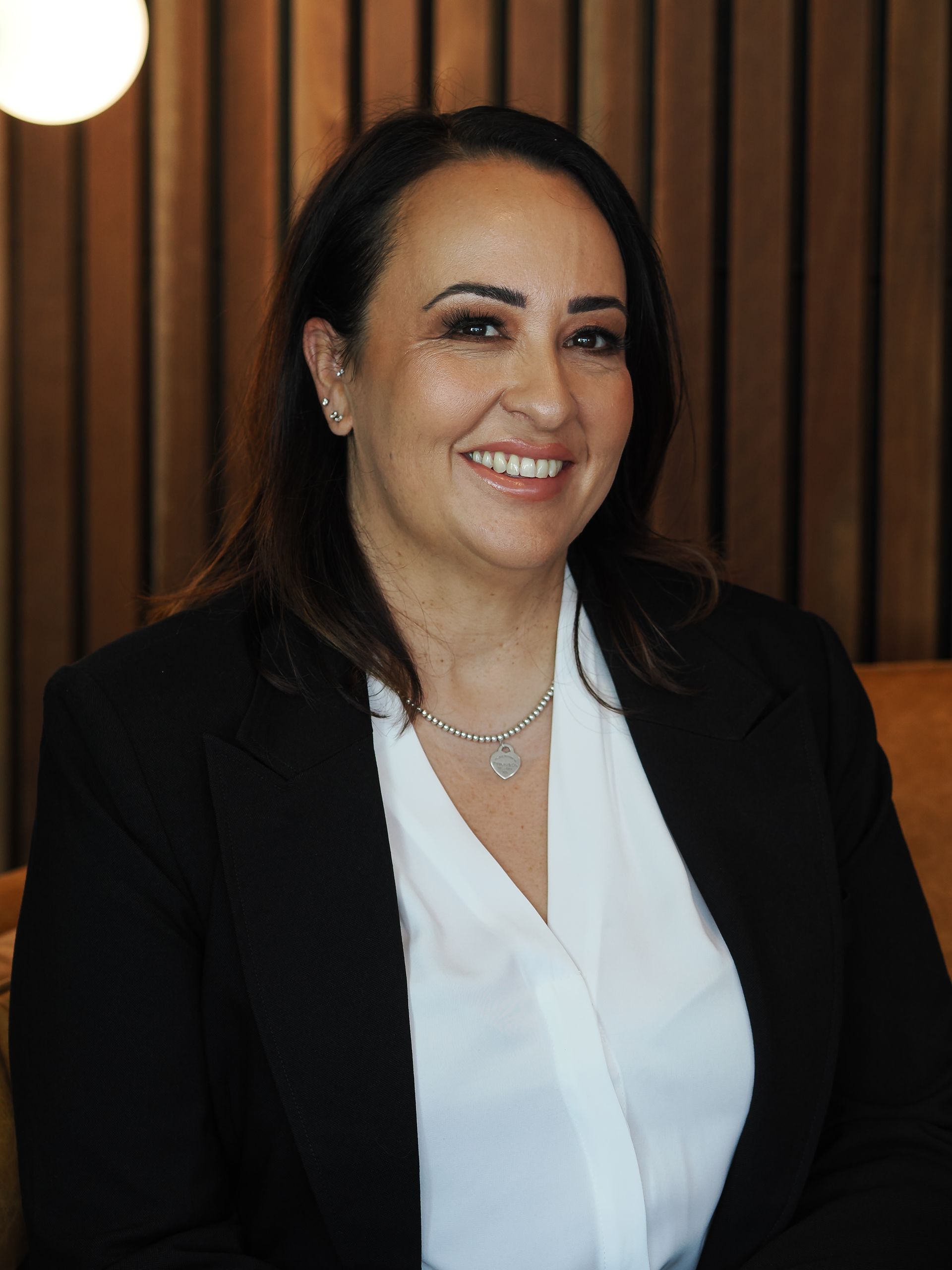 Woman with dark hair smiles while wearing a black blazer over a white shirt; the background has brown paneling.