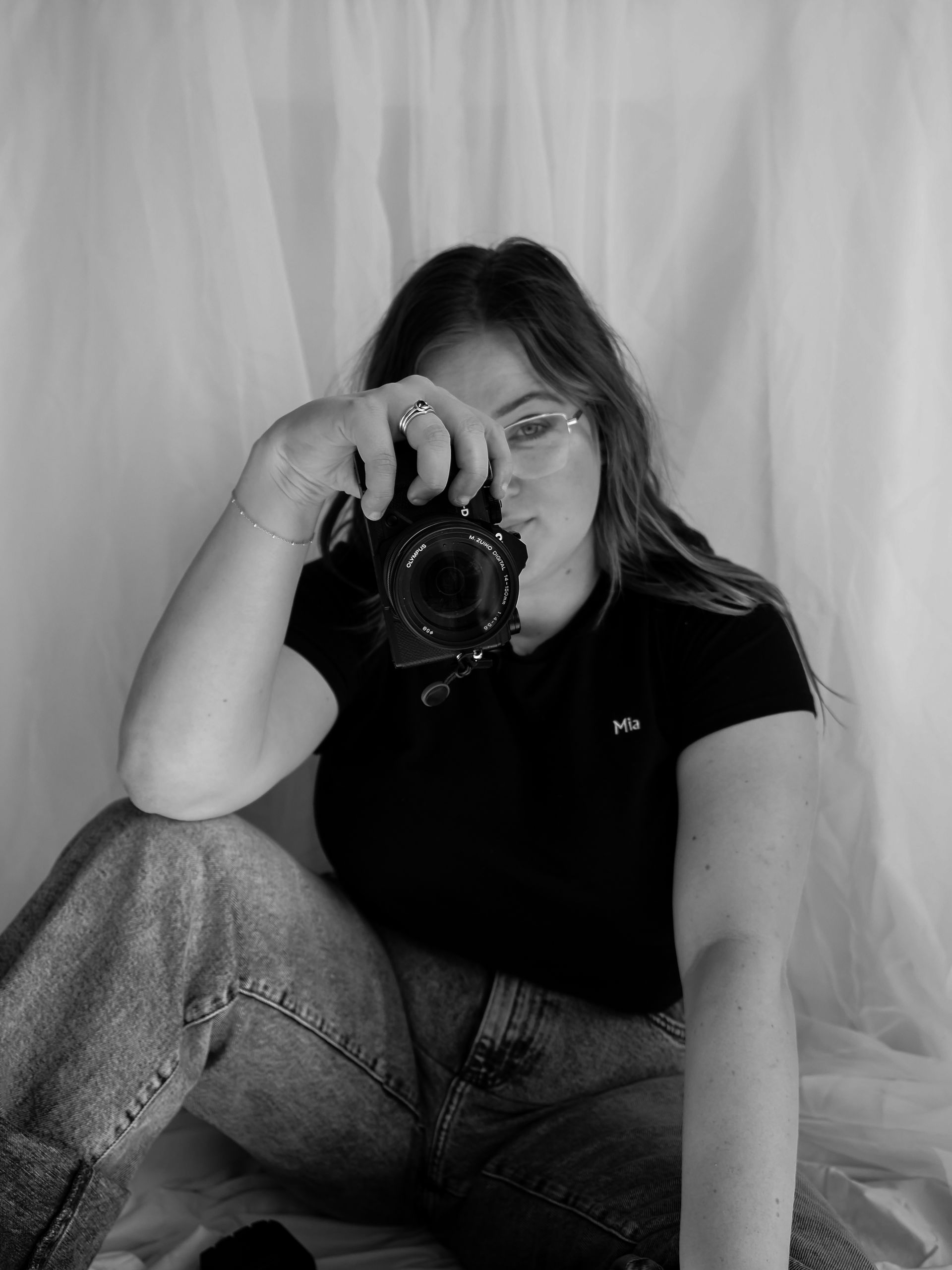 Woman in black shirt and jeans, holding a camera, looking directly at the viewer. White backdrop.